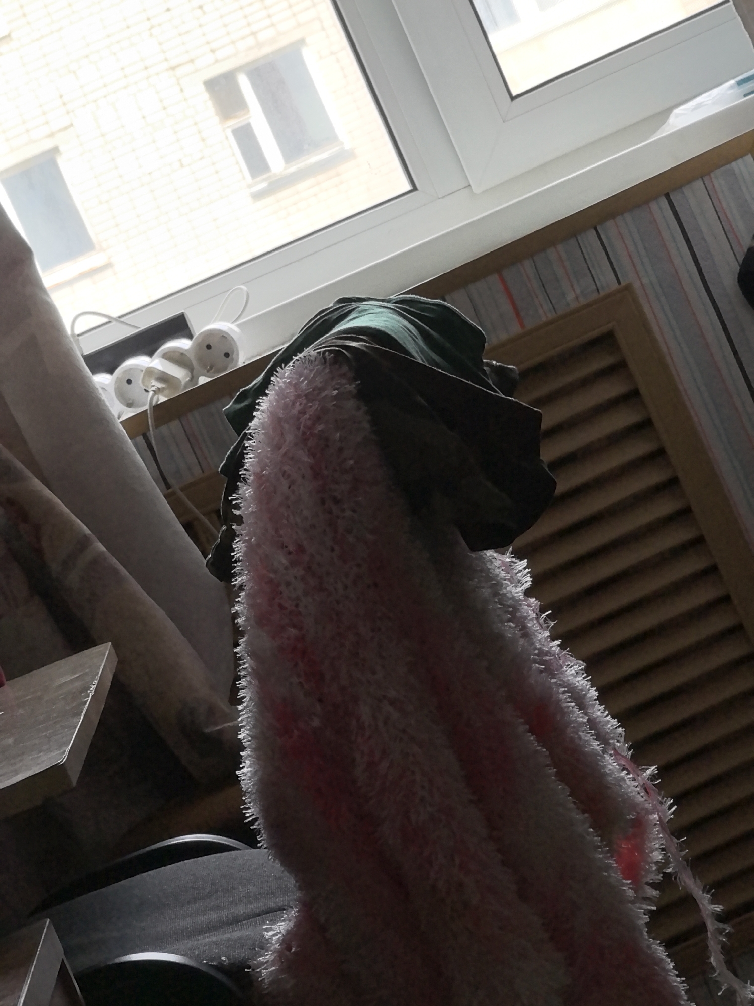 The image depicts an interior scene looking towards a window. Outside the window, a brick building with two rectangular windows is visible. The window in the foreground is a white-framed casement window. On the interior windowsill, several white objects resembling electrical adapters with small circular faces and loops are arranged. A piece of green fabric is draped over the adapters, and a fluffy pink and white textile hangs down in front of the scene. To the right of the green fabric, a wooden louvered door or cabinet is visible. The room appears to be dimly lit.