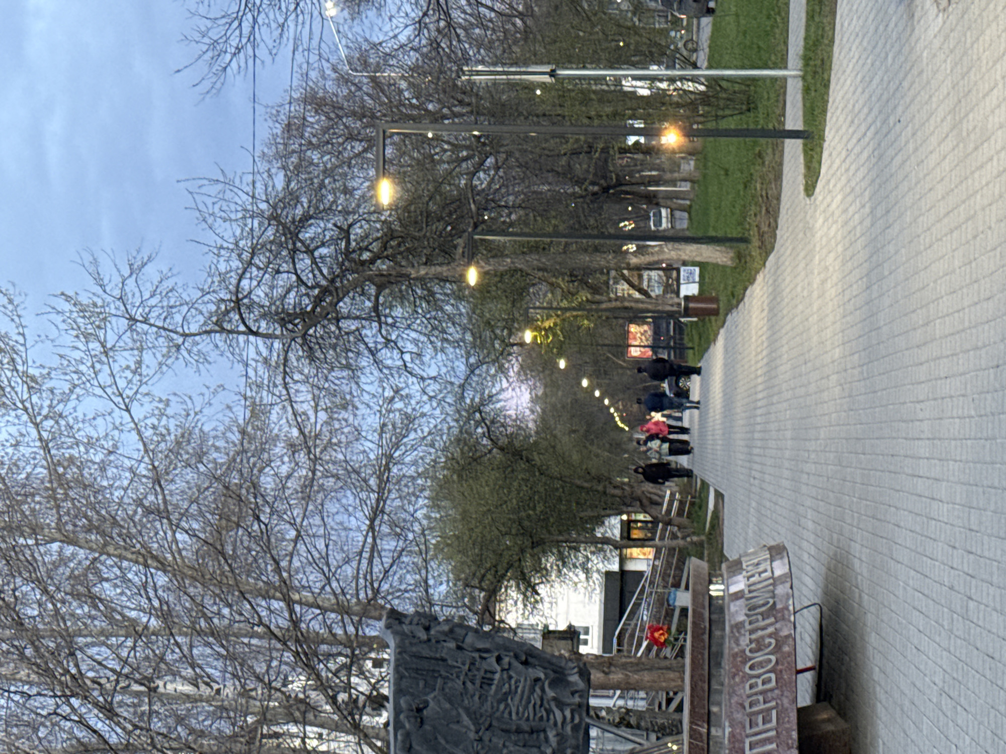 The image captures a tranquil evening scene along a paved pathway, likely a boulevard or park promenade. The pathway, made of light-colored interlocking bricks, extends from the foreground into the distance on the right side of the frame. To its left, a strip of green grass separates the path from a line of numerous deciduous trees, which are currently bare, suggesting late autumn, winter, or early spring.
The path is illuminated by a series of modern streetlights or string lights, which emit a warm, yellow glow and recede into the distance, guiding the eye. Several people are visible in the mid-ground and background, walking along the path. Their figures are somewhat indistinct, appearing as dark silhouettes against the ambient light. The sky above is a soft, muted blue, indicating either dusk, dawn, or an overcast daytime.
In the immediate foreground, on the left side of the path, stands a dark stone monument or plaque. It features a detailed relief sculpture of figures and structures. Below this relief, Cyrillic text is clearly visible, reading "ПЕРВОСТРОИТЕЛЯМ". This translates to "To the First Builders" or "To the Pioneers". The scene evokes a calm atmosphere, characteristic of an evening stroll in an urban or semi-urban setting in Chaikovsky, Russia.