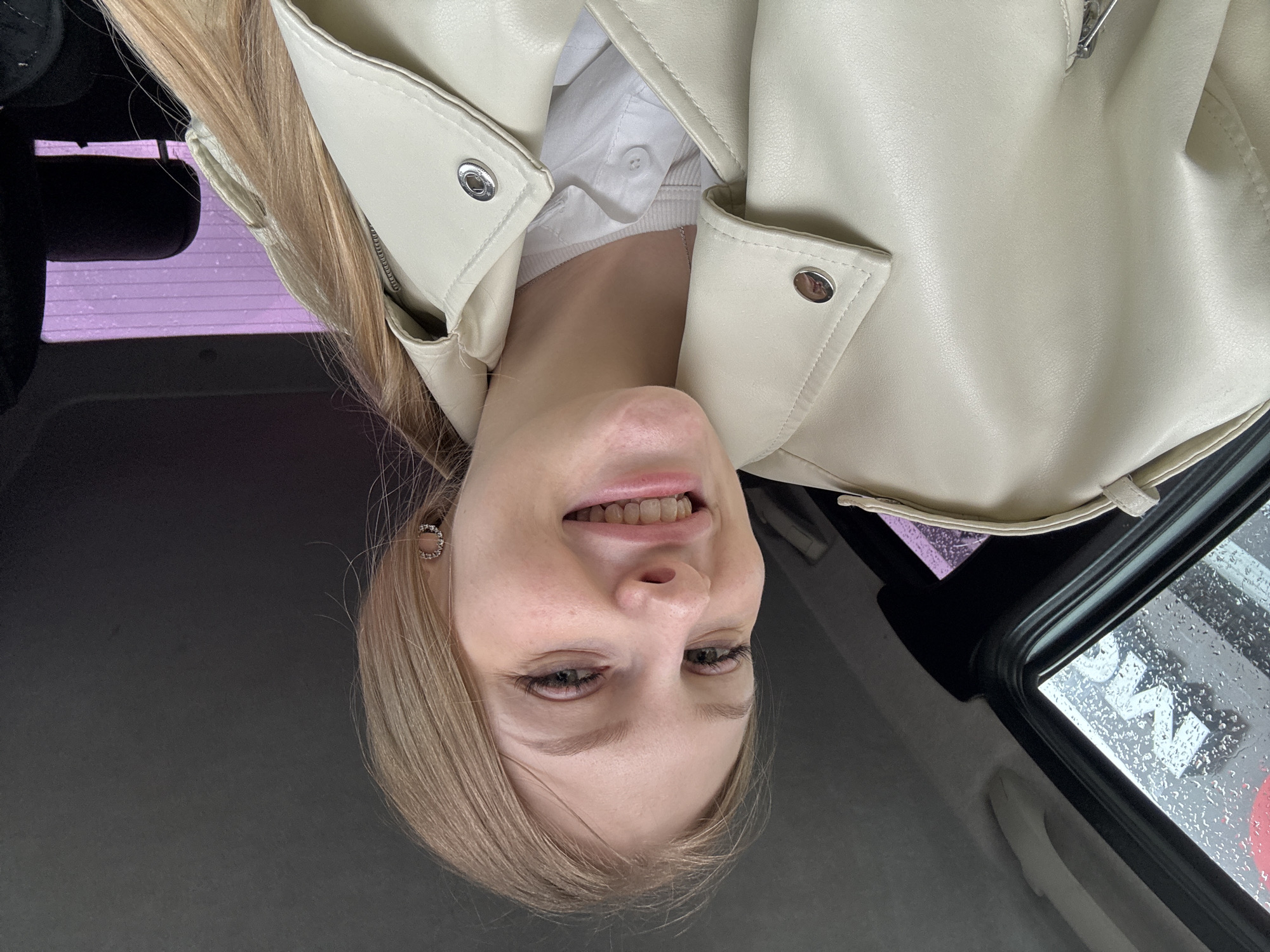 A woman is inside a car in Perm, Russia. The image shows her from a low angle, and the camera is rotated. She has blonde hair and is wearing a light-colored leather jacket over a white shirt. The car window is visible, with rain droplets on the glass. The car's interior is dark. There is no other information available to identify the specific location.