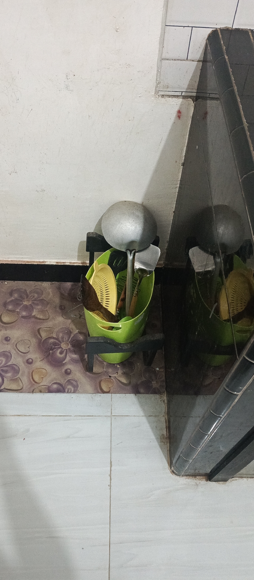 The image depicts a corner of an indoor space, possibly a kitchen or utility area, with a focus on a green plastic bucket filled with kitchen utensils. The bucket is supported by a dark, possibly metal, stand and sits on a floor covered with a patterned mat. The mat features floral designs in shades of purple, brown, and beige. The background consists of a plain white wall that shows some signs of wear and discoloration. To the right of the bucket, a dark, reflective surface, likely a cabinet or appliance, creates a mirrored image of the scene. The floor tiles visible at the bottom of the image are white and appear to be in a grid pattern. The overall lighting suggests an indoor environment with artificial light casting shadows. The scene is static and appears to be an undisturbed arrangement of household items. Given the location information, this scene is situated in Arjasa, Indonesia.