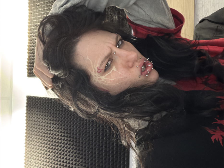 An individual is depicted in a slightly angled, head-and-shoulders indoor portrait. The subject has long, dark, wavy hair, with their right hand positioned within the hair above the ear. The visible left side of the face features elaborate light-colored, web-like markings extending from the temple towards the eye and cheek, with two red crescent shapes above the left eye. Facial piercings include two horizontal barbells below the left eyebrow, two barbells on the bridge of the nose, and numerous spiked studs piercing both the upper and lower lips. The subject wears layered clothing consisting of a dark top with a red graphic on the left chest, a red garment visible underneath, and a light grey long-sleeved top. The background is composed of dark, pyramidal acoustic foam panels covering a wall, indicative of a sound-treated space such as a studio. Overhead lighting illuminates the scene. No specific geographical identifiers are present in the image to determine a location in Sofia, Bulgaria.