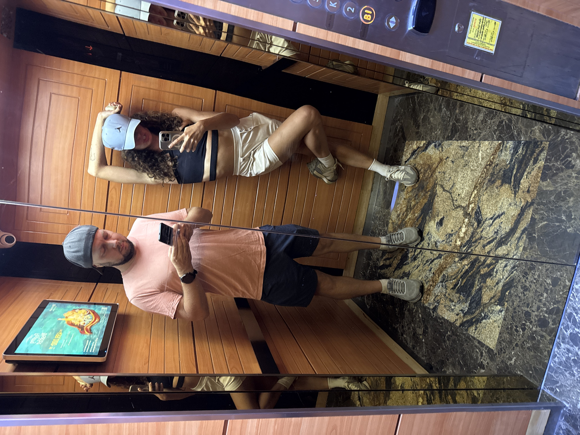 Two people are in an elevator with mirrored walls. A woman with curly hair, wearing a blue baseball cap, a black crop top, and white shorts is lying on the wooden wall of the elevator. She is holding a phone. A man, wearing a gray cap, a pink t-shirt, and blue shorts, is standing. He is also holding a phone. The elevator has a marble floor and a panel with floor buttons. A digital display screen is mounted to the wooden wall. The location is Đà Nẵng, Vietnam.