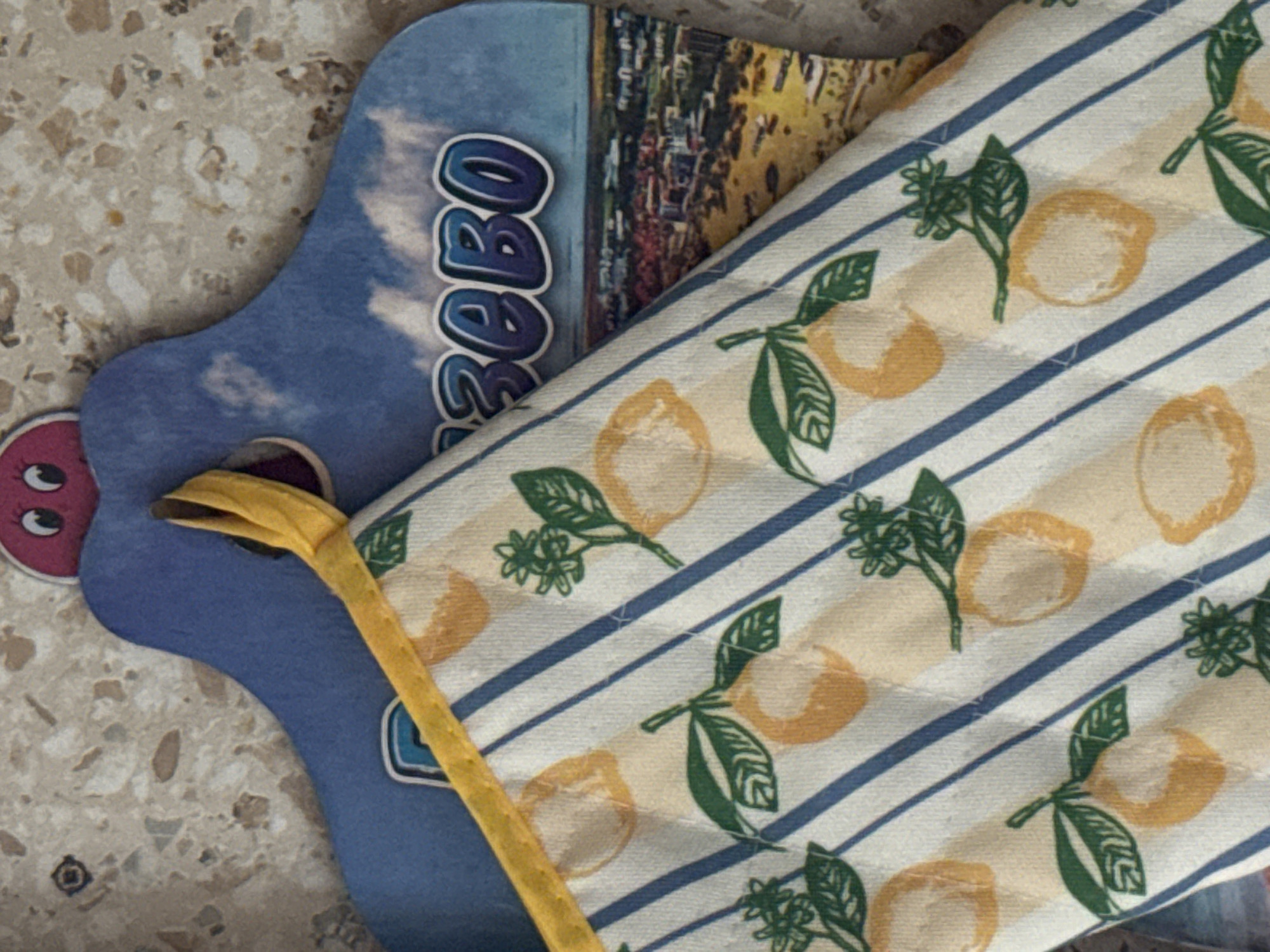 A close-up shot shows a kitchen trivet with a lemon pattern, partially obscuring a souvenir postcard featuring a city skyline and the text "ИЖЕВО" in blue letters. The postcard is placed on a textured, speckled surface.