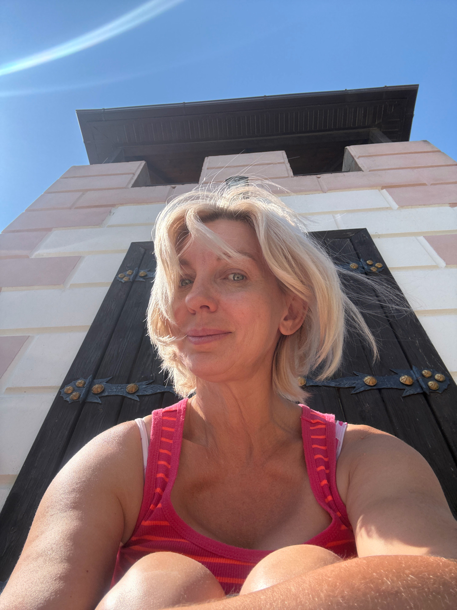 The image captures a woman with blonde hair and fair skin, looking directly at the camera. She is wearing a pink and orange striped tank top, with a white bra strap visible. Her arms and shoulders are visible, with noticeable hair on her right arm. The background features a portion of a building with light pink and white stone blocks, and a large dark wooden door with ornate metal hinges and studs. Above the door, there's a dark roof structure. The sky is clear and blue, with a bright sun creating a lens flare effect. The perspective is from a low angle, looking up at the woman and the building. Based on external information, the building is identified as a guard tower located in the city of Abrau-Dyurso, Russia. The woman is sitting, with her legs crossed and arms resting on her lap, suggesting a relaxed pose. The scene appears to be outdoors on a sunny day.