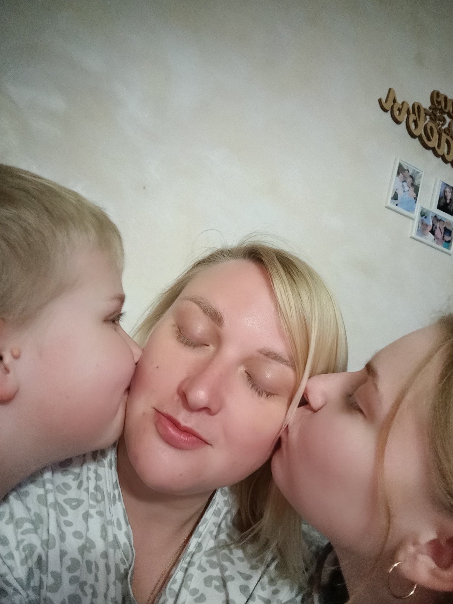 The image shows a selfie of a light-skinned woman with blonde hair being kissed on each cheek by two younger light-skinned individuals. The person on the left appears to be a young child with blonde hair, while the person on the right appears to be a young woman, also with blonde hair. The woman in the center has her eyes closed and appears to be smiling slightly. She is wearing a patterned shirt and a necklace. The background is a light-colored wall with wooden lettering and framed photographs hanging on it. The location is Chaikovsky, Russia.