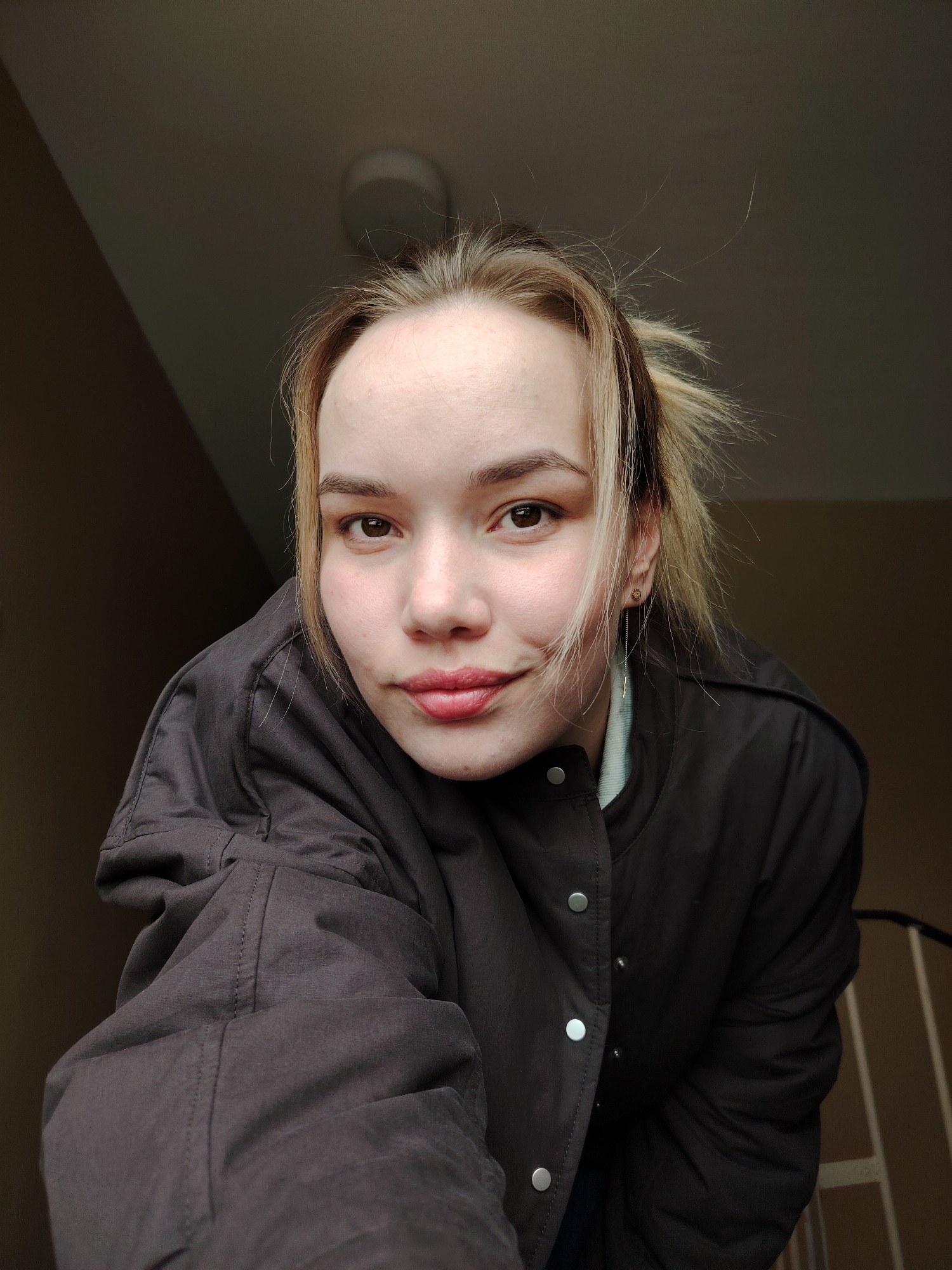 A young adult female is depicted in a close-up, chest-up photographic capture, positioned looking directly into the camera. She has fair skin, brown eyes, and light-toned hair with blonde highlights, with some strands partially obscuring her face. She is dressed in a dark-colored jacket with silver snap buttons, worn over a light-colored inner garment visible at the collar. A small stud earring and a long threader-style earring are visible in her right ear. The background is dim, featuring a dark upper section with a faintly visible ceiling light fixture in the upper right quadrant. A lighter, beige-colored wall segment is present on the mid-right, with vertical and horizontal elements consistent with an interior railing or stairwell visible in the lower right. A diagonal shadow line is cast across the upper left of the frame. The scene is located in the city of Марковский, Russia.