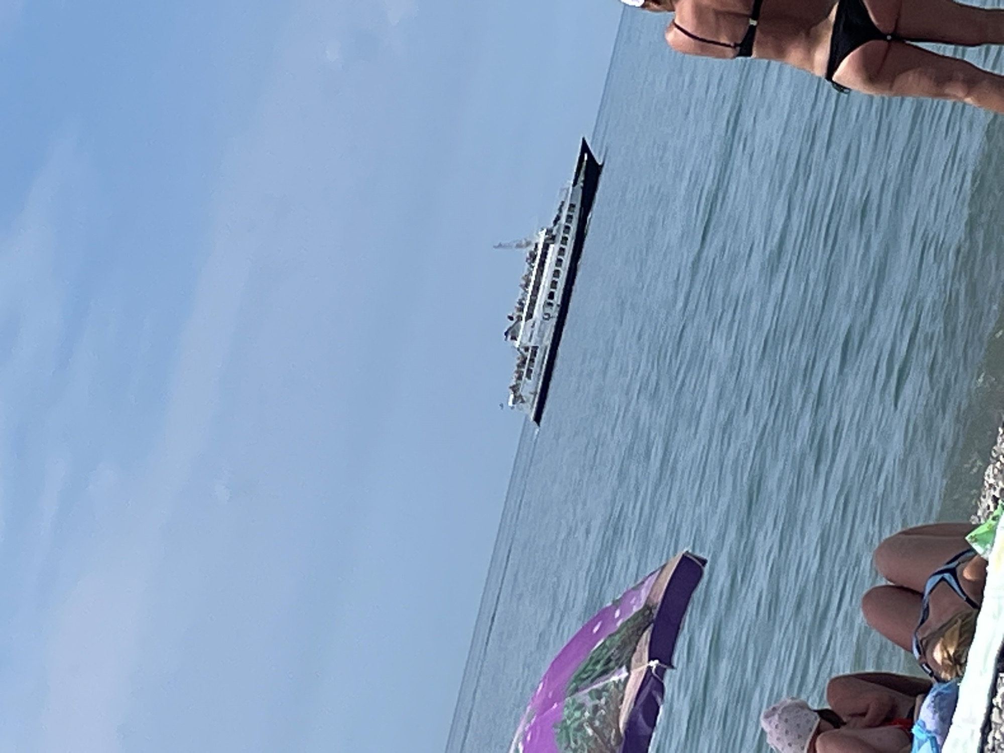 A ferry boat with passengers is visible on the water, approaching from the right of the frame. The boat is white with a dark hull and has multiple decks. In the foreground, on a pebble beach, a person wearing a white cap and a patterned swimsuit is seated, facing away from the camera. To the right of this person, another individual is standing, wearing a black bikini and a white cap. A partially visible purple umbrella with a palm tree design is in the lower left corner of the image. The water is blue and calm, and the sky is clear and bright blue with some wispy clouds.