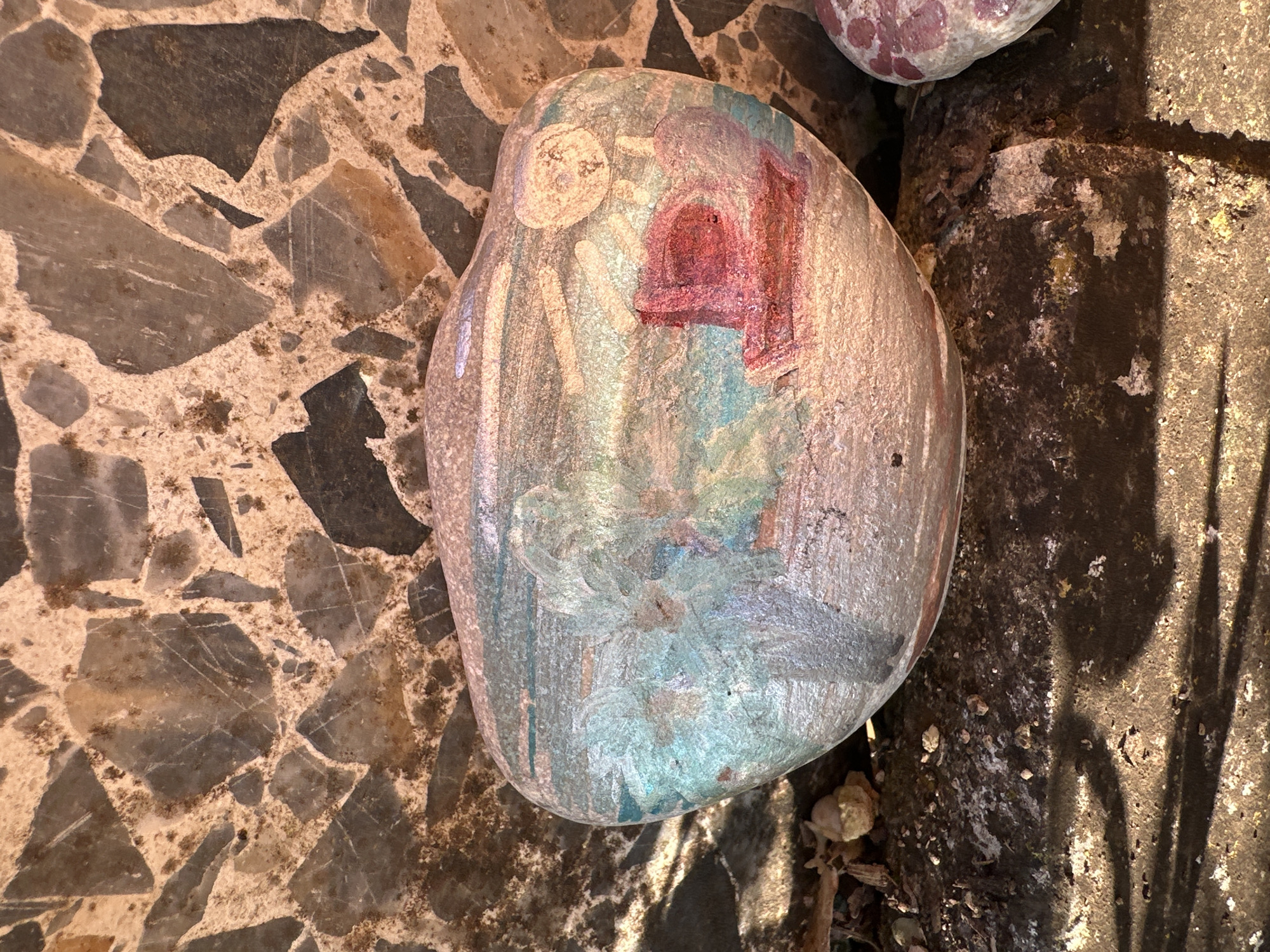 An irregularly shaped, light-colored rock with a weathered painting rests on a mosaic or terrazzo-like ground. The painting on the rock features abstract or stylized forms in muted tones of light blue, teal, white, and purple. A prominent dark red/burgundy shape, possibly depicting a small house or building with an arched doorway, is visible near the top right of the painted area. To its left, a lighter, round shape suggests a head or celestial body.
The ground beneath the rock is composed of irregularly shaped, dark grey and brown stones set within a lighter, sandy-colored matrix. To the right of the main painted rock, a dark, textured surface, possibly concrete or compacted earth, is visible. Long, slender shadows are cast across this dark surface, indicating natural sunlight and nearby objects or foliage. Part of another painted rock is visible in the upper right corner of the image. The scene is outdoors, likely during the daytime in Sesto Campano, Italy. There is no discernible text.