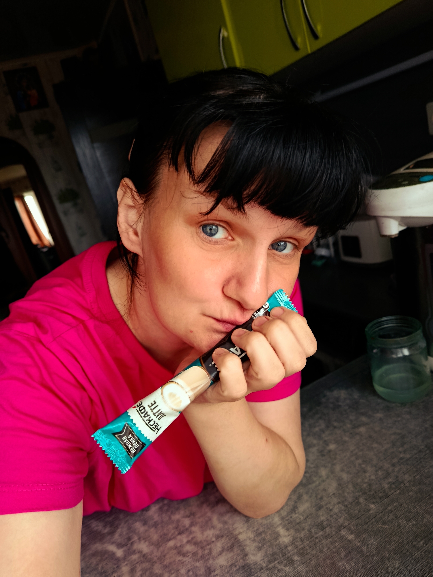 A woman with short black hair and blue eyes, wearing a pink T-shirt, is indoors, holding a single-use "Nescafe Latte" stick in her hand. The text "НЕСКАФЕ ЛАТТЕ НЕЖНАЯ ПЕНКА" is visible on the stick. A glass jar with water is on the table next to her. The location is in Фоки, Russia.