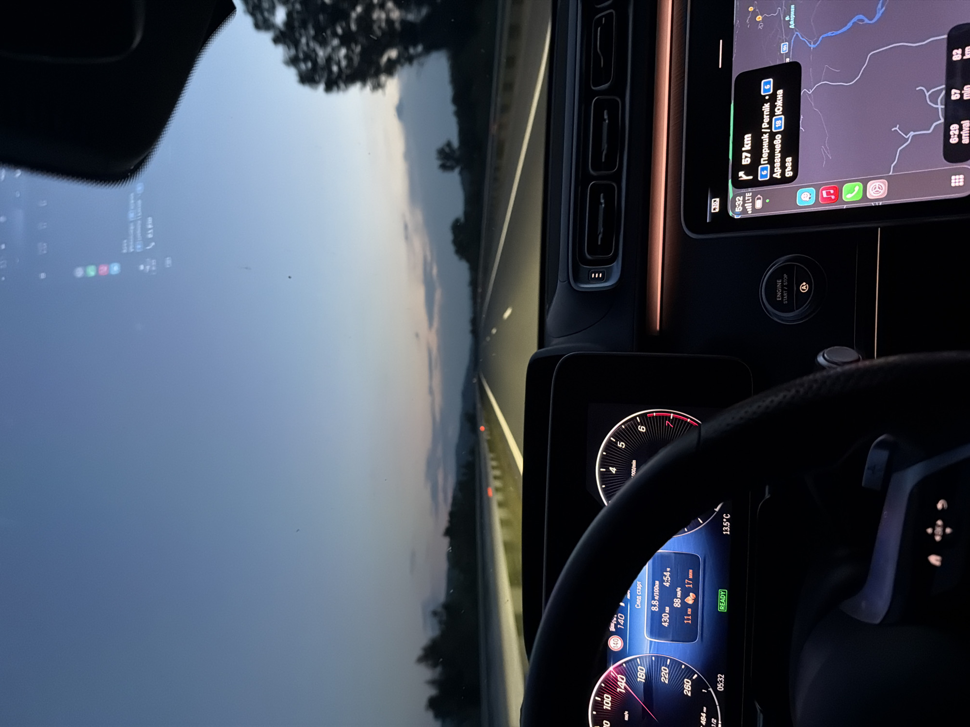 The image shows the interior of a car, focusing on the dashboard and windshield view. The car is in motion on a multi-lane highway at twilight. The speedometer reads about 140 km/h. The infotainment system displays data regarding the trip, showing 430 kilometers driven, with a fuel consumption of 8.8 liters per 100 km. The outside temperature is 13.5 degrees Celsius. A smartphone mounted near the dashboard is displaying a navigation app, showing the destination as Pernik, Bulgaria, 57 km away, accessible via road 6. Given the destination and the "Южна дъга" mentioned on the screen, it is unlikely the location is Mursalevo.