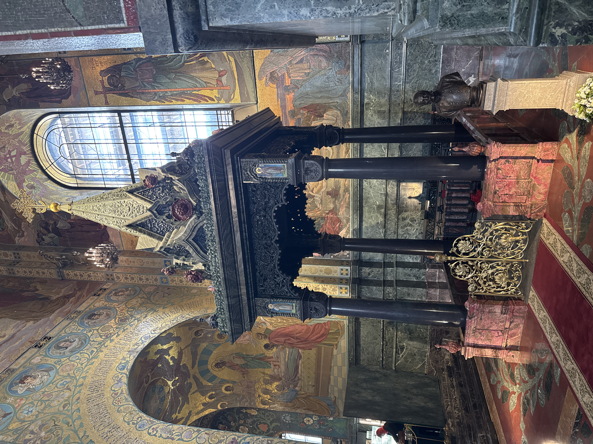 The image captures the interior of the Church of the Savior on Spilled Blood in Saint Petersburg, Russia. The ornate structure in the foreground is a dark, intricately carved canopy supported by four black pillars. Above the canopy is a large arched window, through which daylight streams in. The walls and ceilings are covered in mosaics depicting religious figures and scenes. A bronze bust sits atop a marble pedestal to the right of the canopy. A red carpet with a patterned border extends towards the right edge of the image. The flooring is also decorated with intricate patterns. Chandeliers hang from the ceiling.