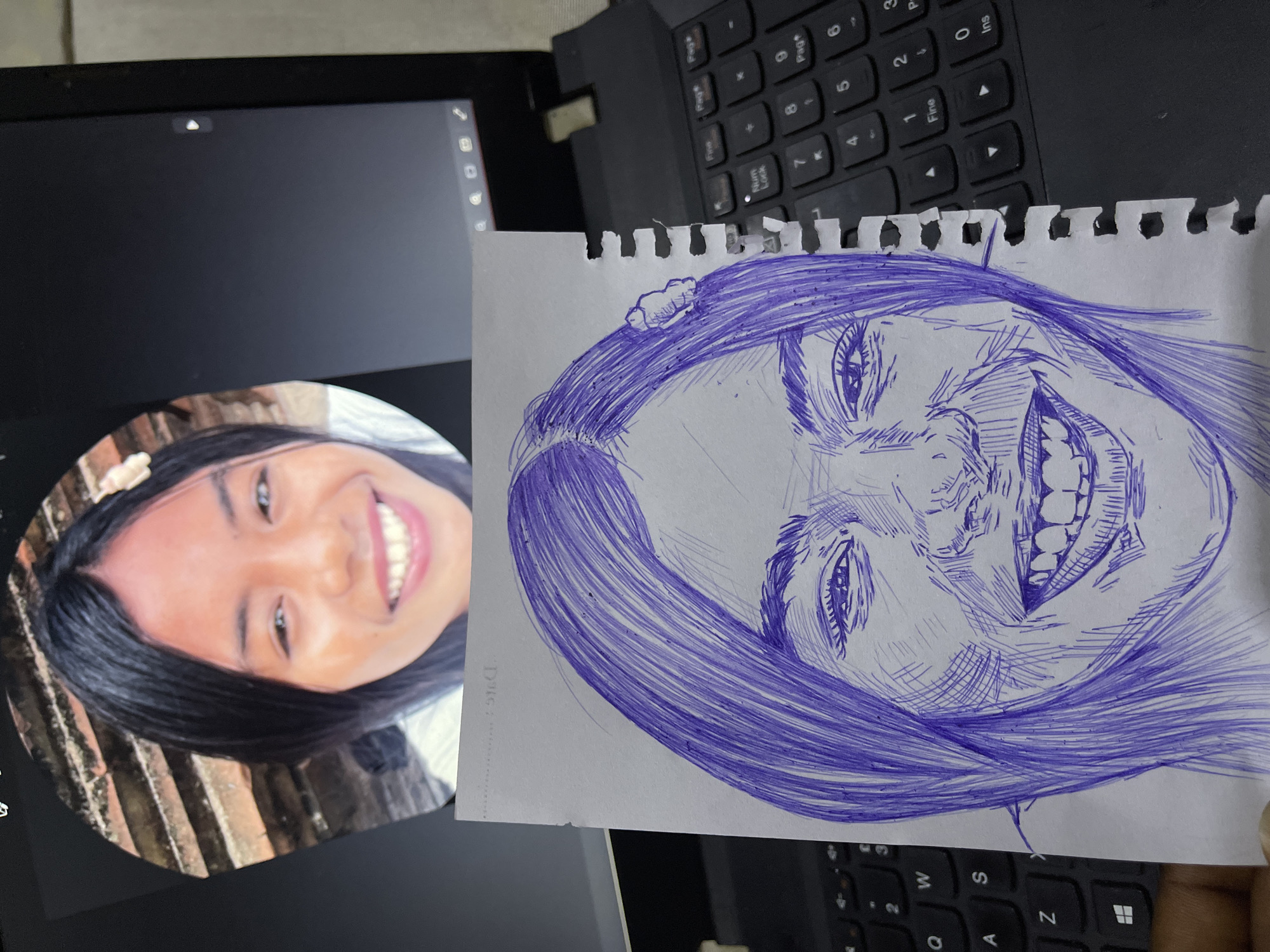 A close-up, eye-level shot shows a laptop with a picture of a young woman on its screen. Below the laptop, a hand holds a sketch of the same woman, drawn in blue ink on white paper. The sketch is a close-up of her face, emphasizing her smile and hair. The laptop's keyboard is visible in the foreground, and the overall scene appears to be indoors. There is no indication of the location being in Abuja, Nigeria.