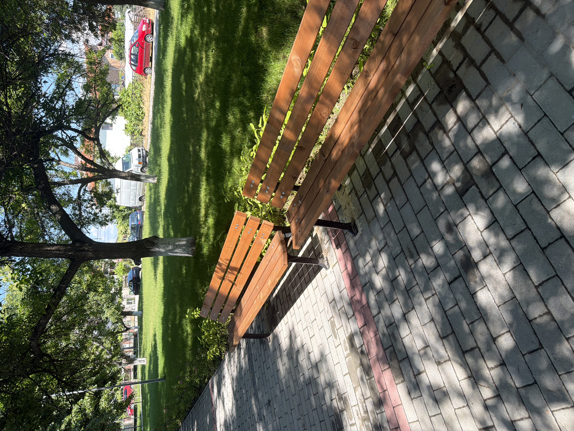 The image shows a paved walkway with two wooden park benches. Beyond the walkway is a well-maintained lawn with several trees. In the background, there are houses and parked vehicles, including a red car, a white van, and a dark-colored car. The lighting suggests it is daytime, with dappled sunlight filtering through the trees, casting shadows on the walkway and grass. This scene is located in the city of Zlatitrap, Bulgaria.