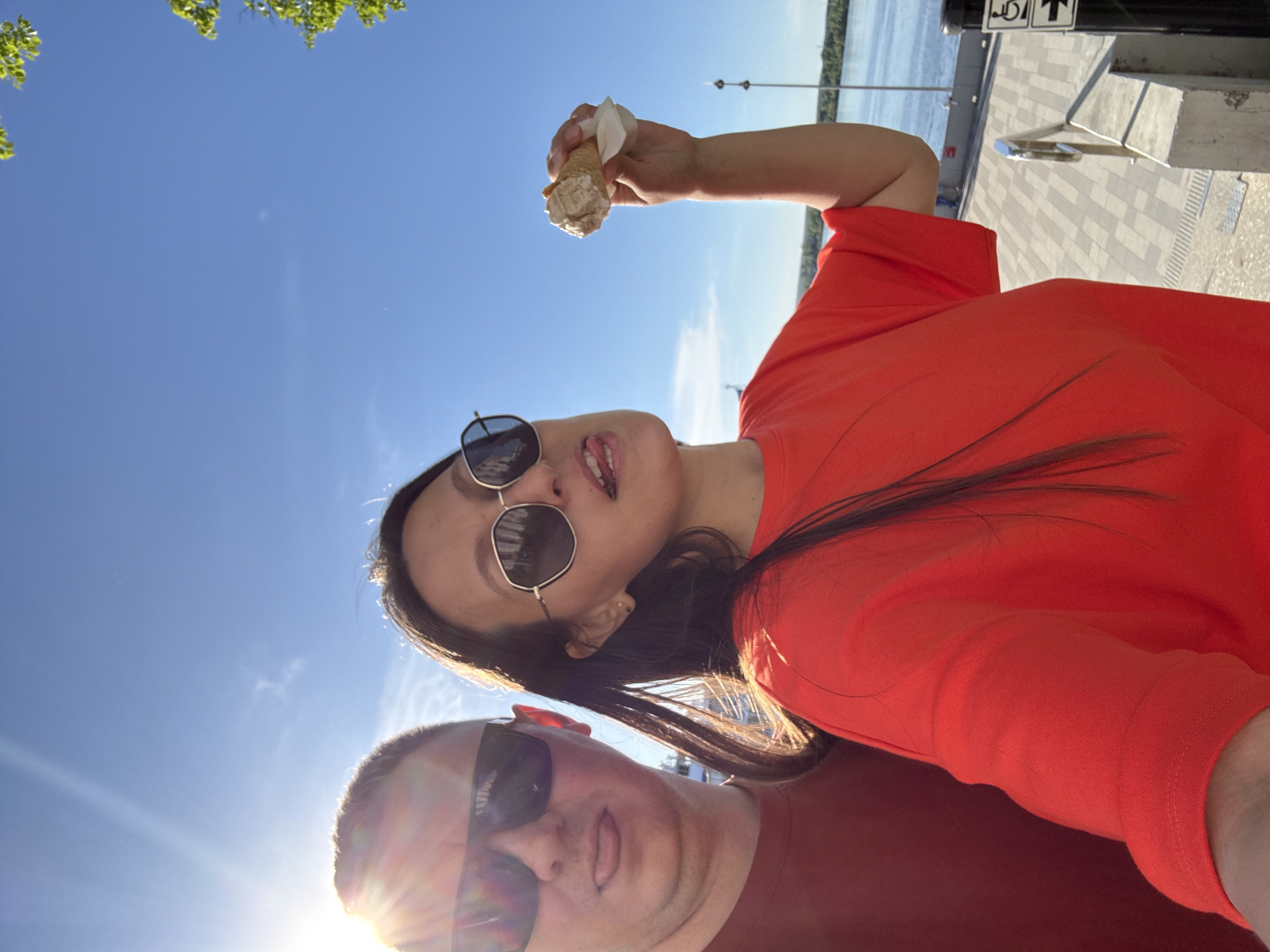 A low-angle selfie shows a woman and a man with an ice cream cone against a bright blue sky. The woman is wearing a coral-colored t-shirt and sunglasses, holding an ice cream cone in her raised right hand. The man, wearing sunglasses and a maroon-colored shirt, is positioned in the lower portion of the frame. A river, trees, and a waterfront walkway with benches are visible in the background. The photograph was taken in the city of Perm, Russia.