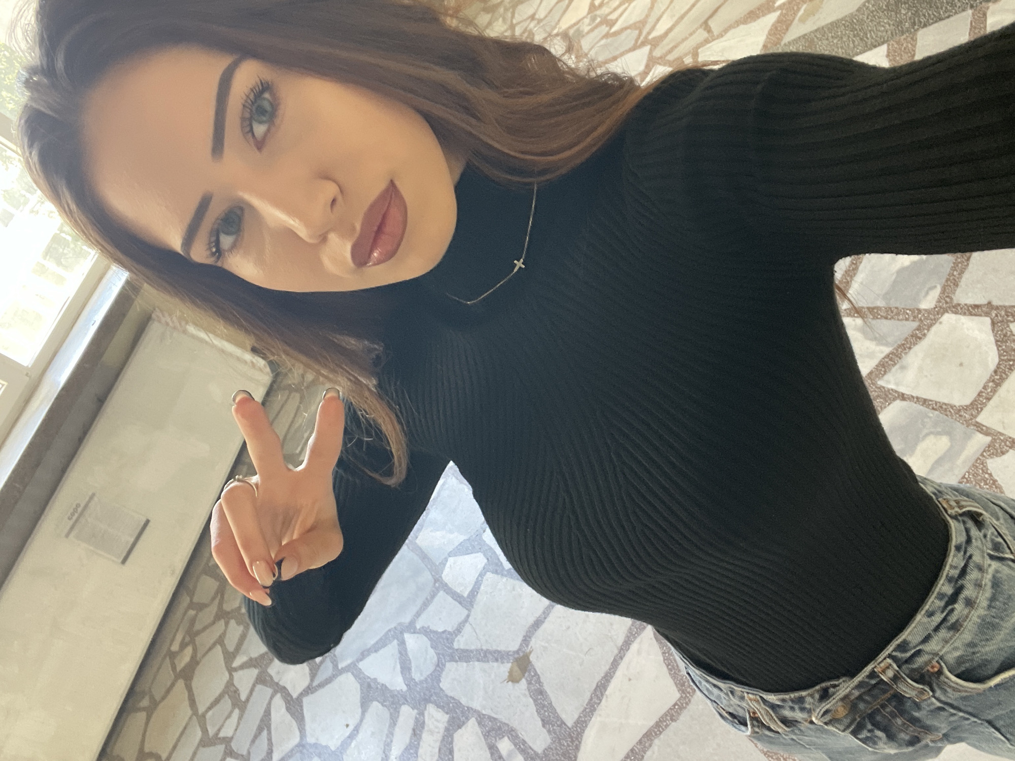 A young woman is taking a selfie indoors. She is wearing a black turtleneck sweater and light blue jeans. She is holding up a peace sign with her left hand. She has brown hair and blue eyes. She is wearing a necklace with a small cross pendant. The floor is tiled with a geometric pattern. A window is visible in the background on the left. It is not possible to determine the exact location in Veliko Tarnovo, Bulgaria, from the image alone.