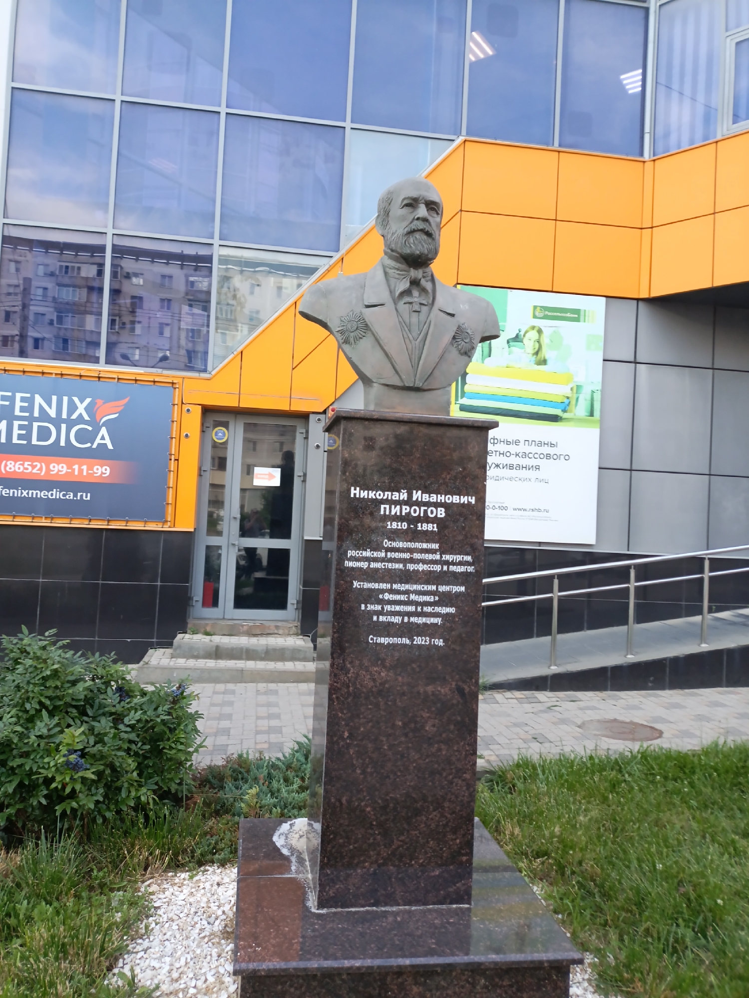 A bust of Nikolai Pirogov stands on a granite pedestal in front of a modern building with orange and grey panels and large blue windows. The bust depicts a bearded man wearing a suit and medals. The pedestal has an inscription in Russian, which translates to: "Nikolai Ivanovich Pirogov, 1810-1881. Founder of Russian military field surgery, anesthesiology, professor and teacher. Installed by the medical center 'Phoenix Medica' as a sign of respect for the legacy and contribution to medicine. Stavropol, 2023." A sign for "Fenix Medica" is visible on the left side of the building, along with a phone number and website. The area around the bust is paved with cobblestones and surrounded by grass and a small bush. The building appears to be a medical facility. The overall scene is a dedication to a prominent historical figure in medicine.