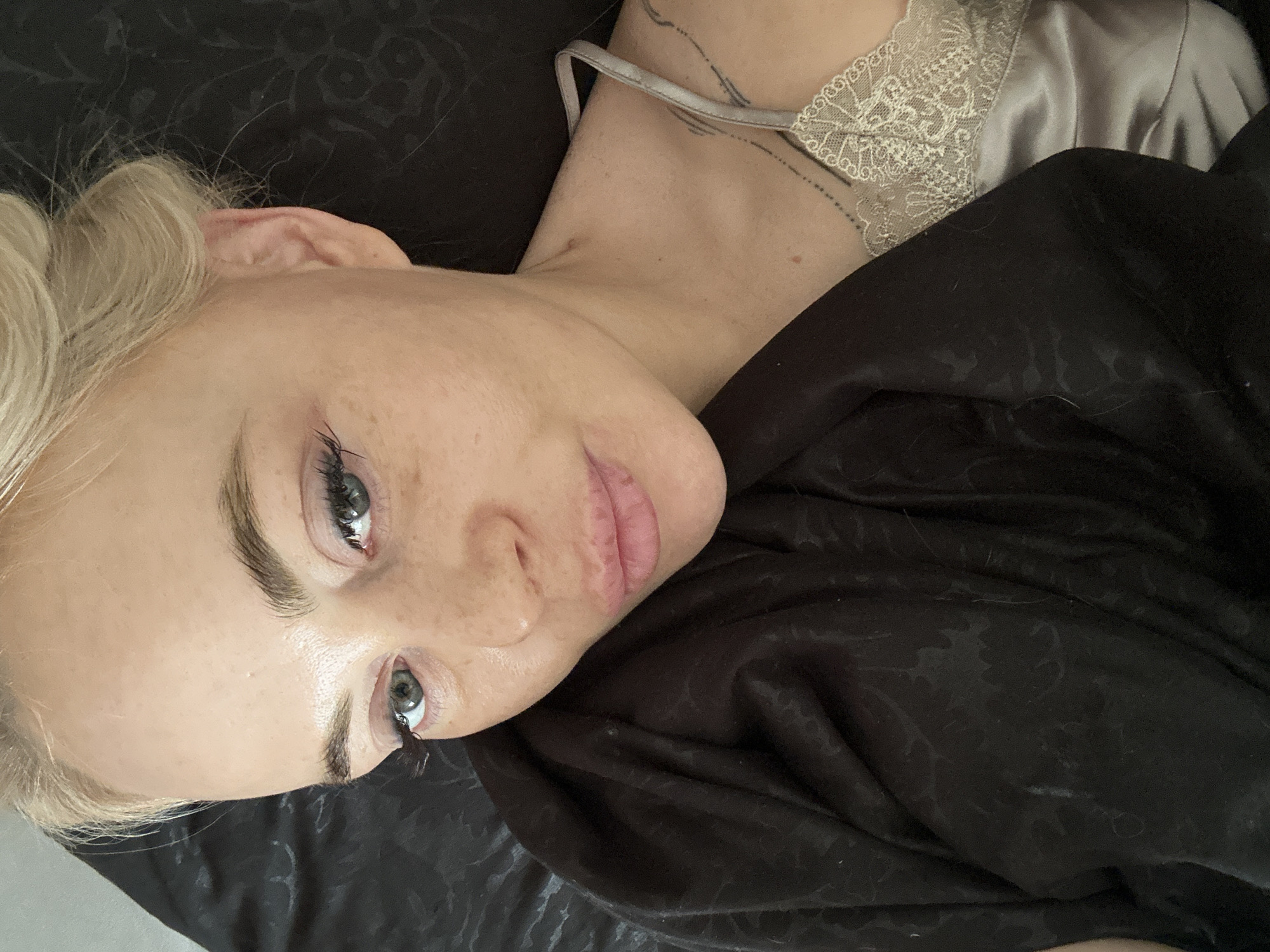 The image depicts an indoor setting in the city of Unknown, Unknown. An adult female with blonde hair, styled in an updo, is lying in a reclined position, looking directly towards the viewer. Her fair complexion shows visible freckles/moles across her nose and cheeks. She has light-colored eyes, appearing greenish-grey, accentuated by dark eyeliner and voluminous eyelashes. Her eyebrows are dark brown and well-defined. Her lips are full, with a light pink hue, possibly from lipstick or gloss.
She wears a light grey or champagne-colored satin camisole with intricate lace trim along the neckline. A dark, possibly black, fabric with a subtle embossed or patterned texture covers her left shoulder and extends across the lower portion of the frame, suggesting a blanket or bedding. A faint linear tattoo, possibly text or symbols, is visible on her left shoulder/upper arm. A thin silver-colored chain necklace with a small pendant is partially visible around her neck. The immediate background is primarily composed of the dark, patterned fabric, suggesting a bed or soft furnishing.