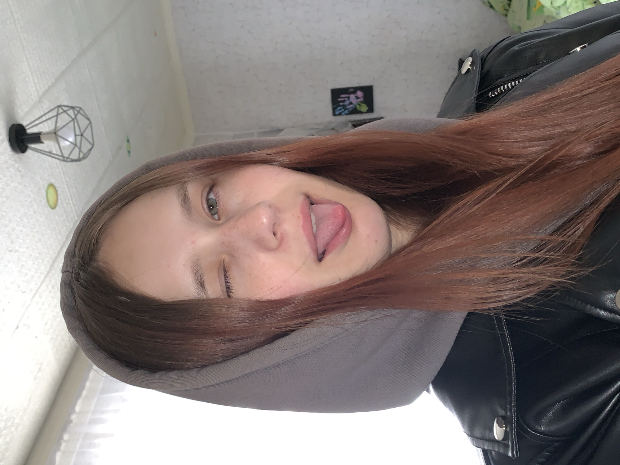 A person with red hair is wearing a grey hoodie and a black leather jacket. They are sticking their tongue out and winking. A lamp and a small framed image are visible in the background. The location is unknown and cannot be determined from the image.