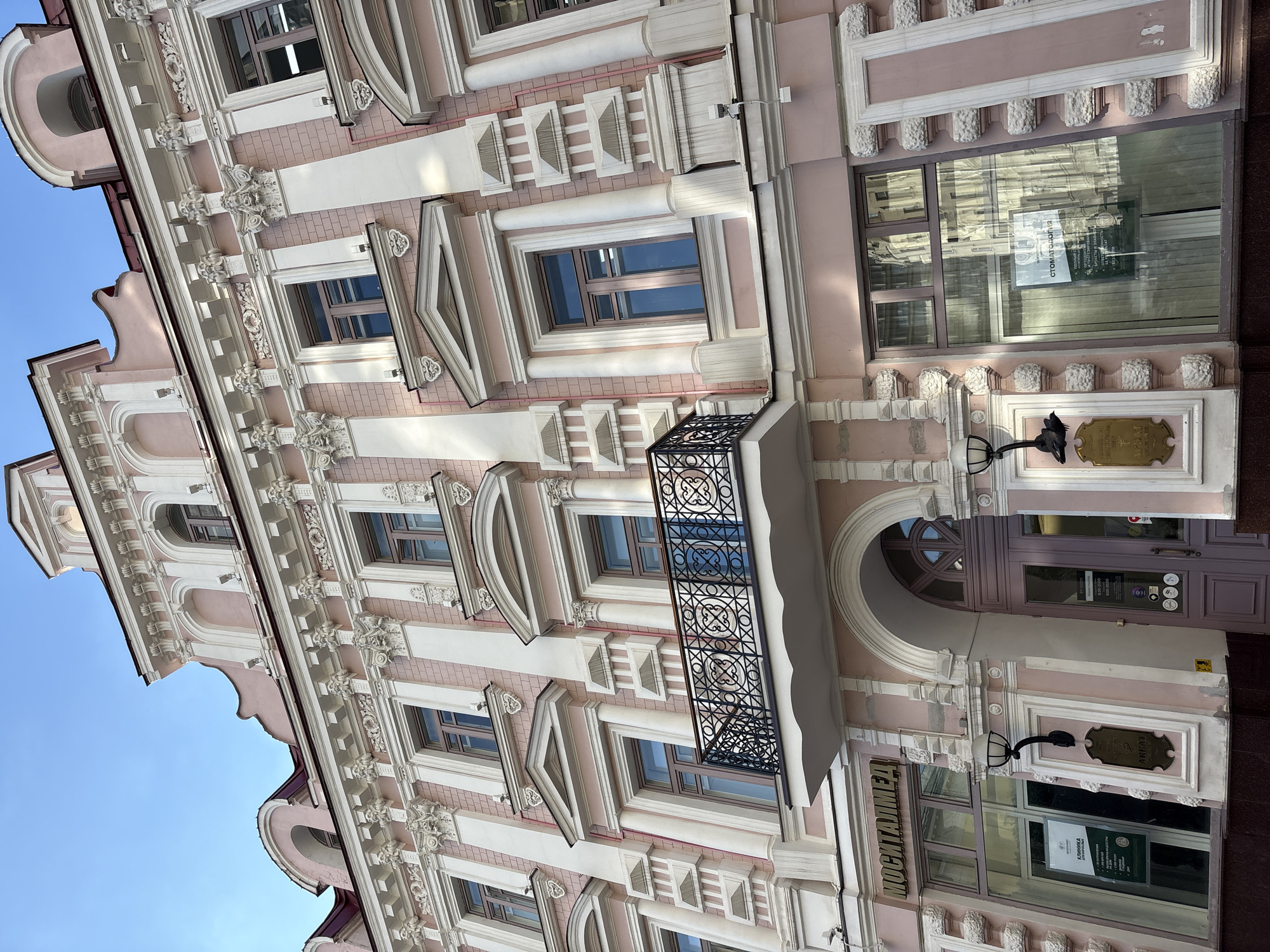 The image shows the exterior of a pink and white building with ornate architectural details, including columns, decorative moldings, and arched windows. There is a balcony with a wrought-iron railing on the second floor. The building appears to be in an urban setting, with a clear blue sky overhead. A sign above a storefront reads "МОСИТАЛМЕД" (MOSITALMED). Another sign on the storefront window says "СТОМАТОЛОГИЯ" (DENTISTRY). The building's design suggests it is likely located in a historic district of Moscow, Russia, possibly with a European architectural influence. No people are visible in the image.