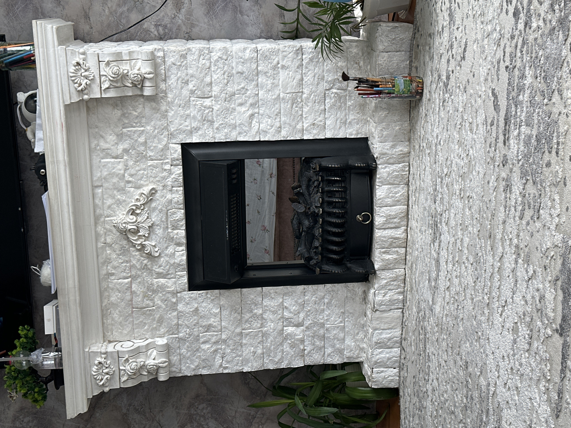 A white stone fireplace with a black electric insert is the central focus of this indoor scene. The fireplace is adorned with decorative carvings on its mantelpiece and sides, featuring floral motifs. To the left of the fireplace, a large potted plant with green leaves stands. To the right, a glass jar filled with art brushes and pencils sits on the hearth. The floor is covered with a light-colored, patterned rug. The background wall has a textured gray finish. On top of the mantelpiece, there are various objects including a small potted plant, a glass bottle with a light, and a white box.