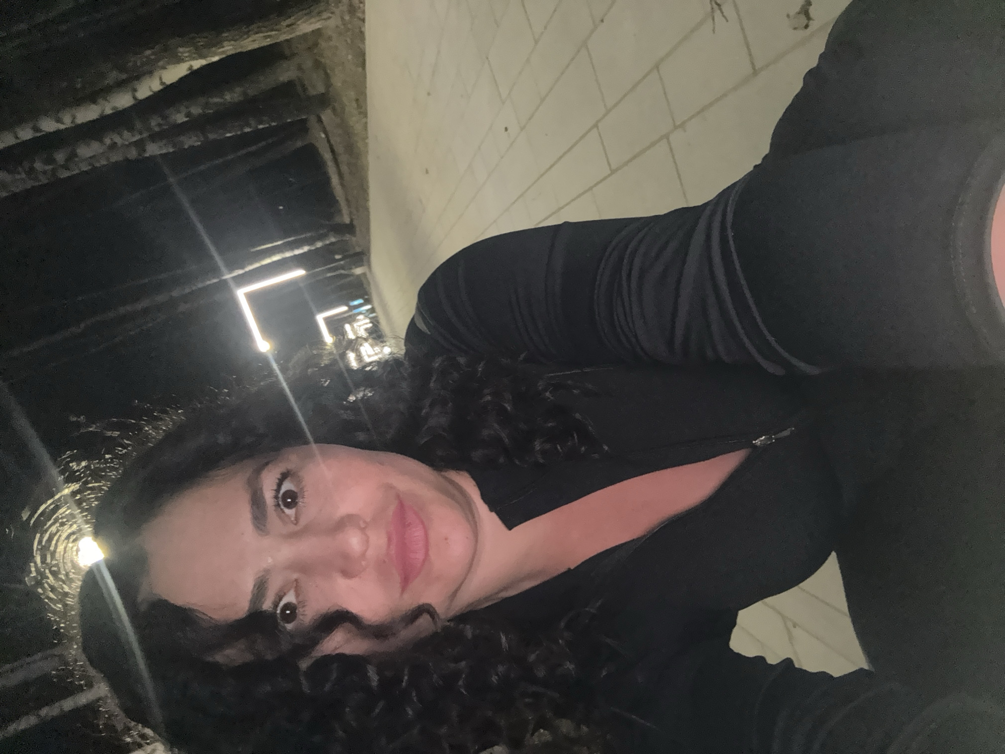 A woman with dark curly hair is taking a selfie in an underpass. The underpass has a ceiling with wooden beams and light fixtures. One of the walls is made of white bricks. The woman is wearing a dark-colored athletic jacket.