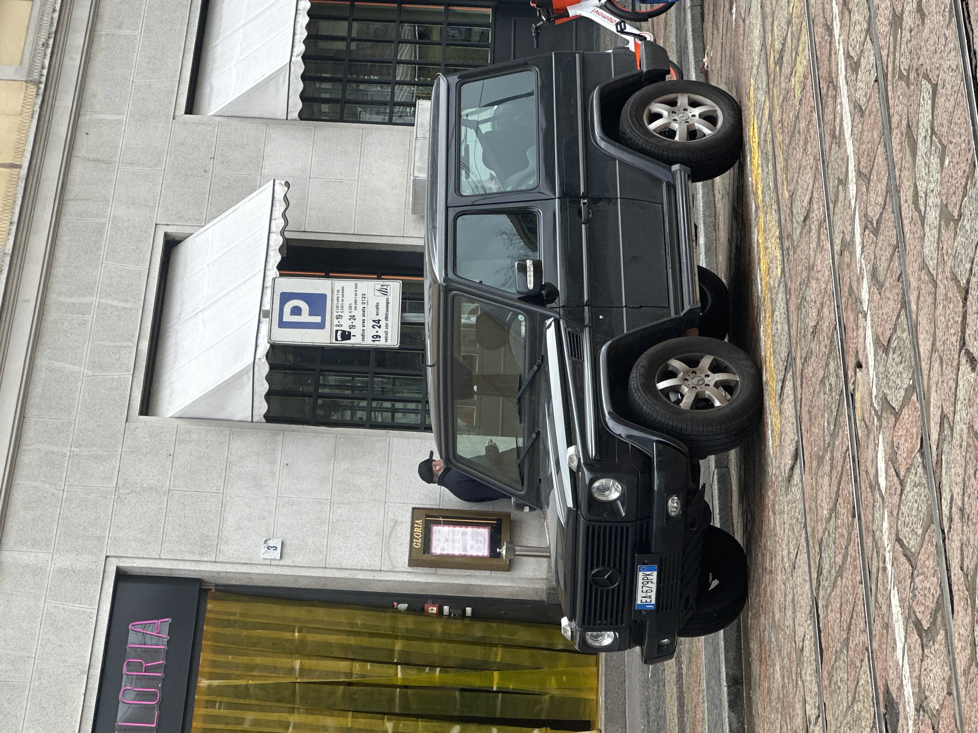 The image captures a daytime street scene at Via San Protaso 3, Milan, Italy, under overcast lighting conditions.
A black Mercedes-Benz G-Class, a two-door variant, is parked on a reddish-brown cobblestone street. The vehicle displays an Italian license plate, "EA 679PK," on its front bumper and features multi-spoke alloy wheels. Two parallel tram tracks are visible on the cobblestone surface, extending through the foreground and beneath the vehicle.
A male individual, dressed in a dark jacket and a dark baseball cap, stands near the front left of the G-Class, facing a wooden-framed menu board labeled "GLORIA" that is mounted on the building facade. A faint, dark silhouette of a person is discernible within the rear passenger compartment of the G-Class.
The building behind is constructed with light grey, rectangular stone blocks. On the left, a window features a bright pink neon sign displaying "LORIA," positioned above vibrant yellow curtains. The civic number "3" is affixed to the facade between this window and an adjacent one. To the right, two windows with dark frames and multiple panes are visible, each topped with a white fabric awning with scalloped edges. A blue rectangular parking sign, indicating regulations for "8-19" and "19-24" hours and marked "codice area sosta 0123," is attached to the wall between the awnings. A white and orange shared bicycle is partially visible on the far right, positioned behind the G-Class. A dark pole with a red button is attached to the building wall to the left of the menu board.