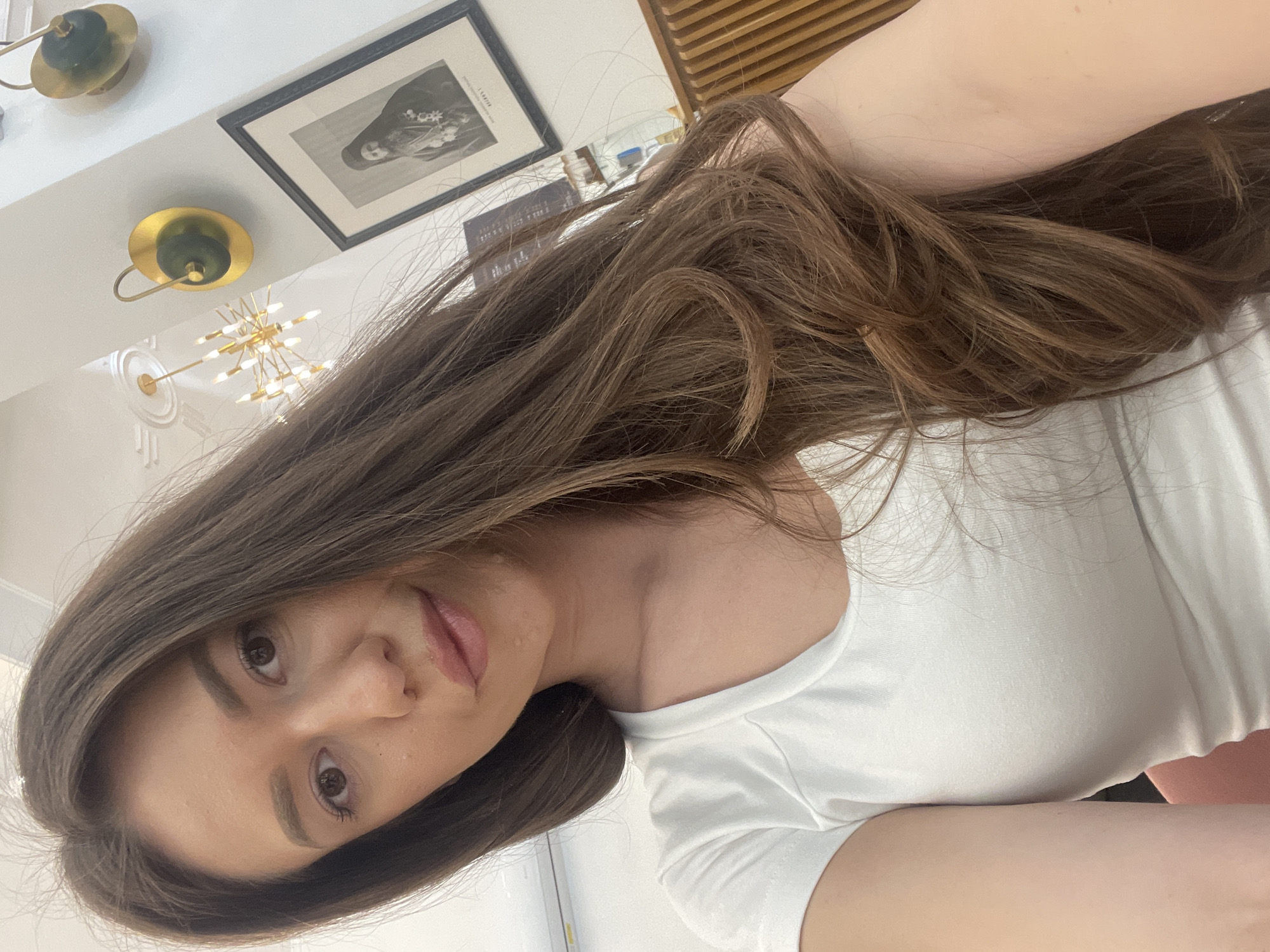A woman with long brown hair and wearing a white t-shirt is facing the camera. The background includes a picture frame, ceiling lights, and a wooden slatted wall. The location is not discernible from the image.