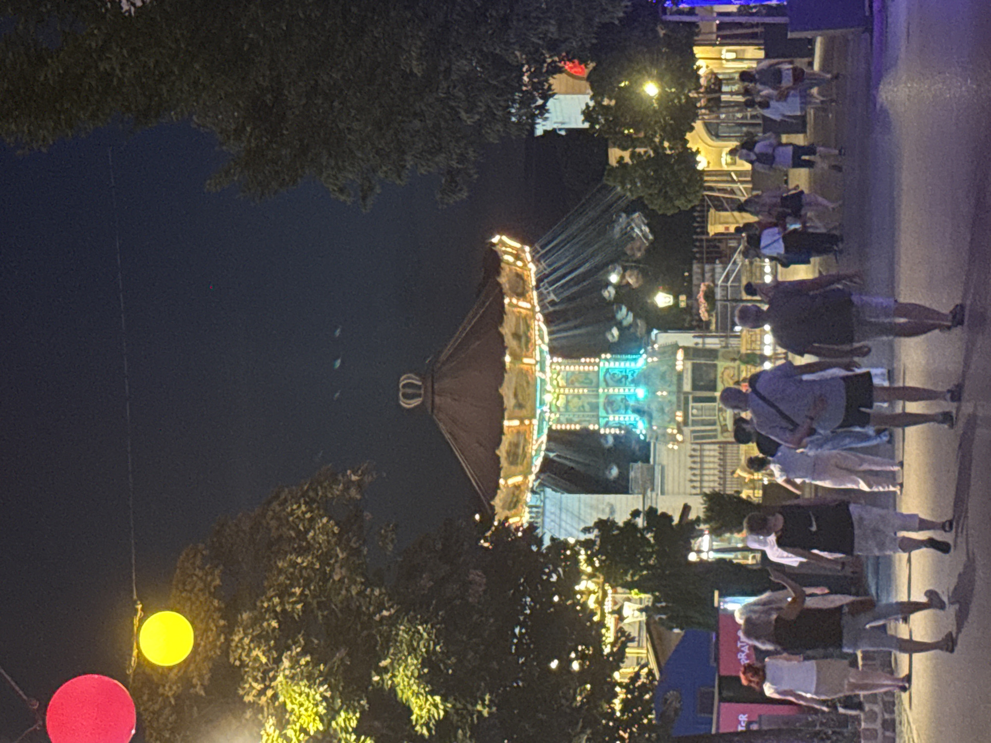 The image captures a scene at night in a lively amusement park, likely located in Vienna, Austria, given the presence of a well-known amusement park. The central focus is a brightly lit carousel, its vibrant lights creating a magical atmosphere against the dark sky. Surrounding the carousel, people are seen walking and enjoying the evening, their silhouettes and forms illuminated by the park's colorful lighting. Trees with illuminated foliage frame the scene, adding to the festive ambiance. The overall impression is one of a cheerful and engaging evening at a popular attraction.