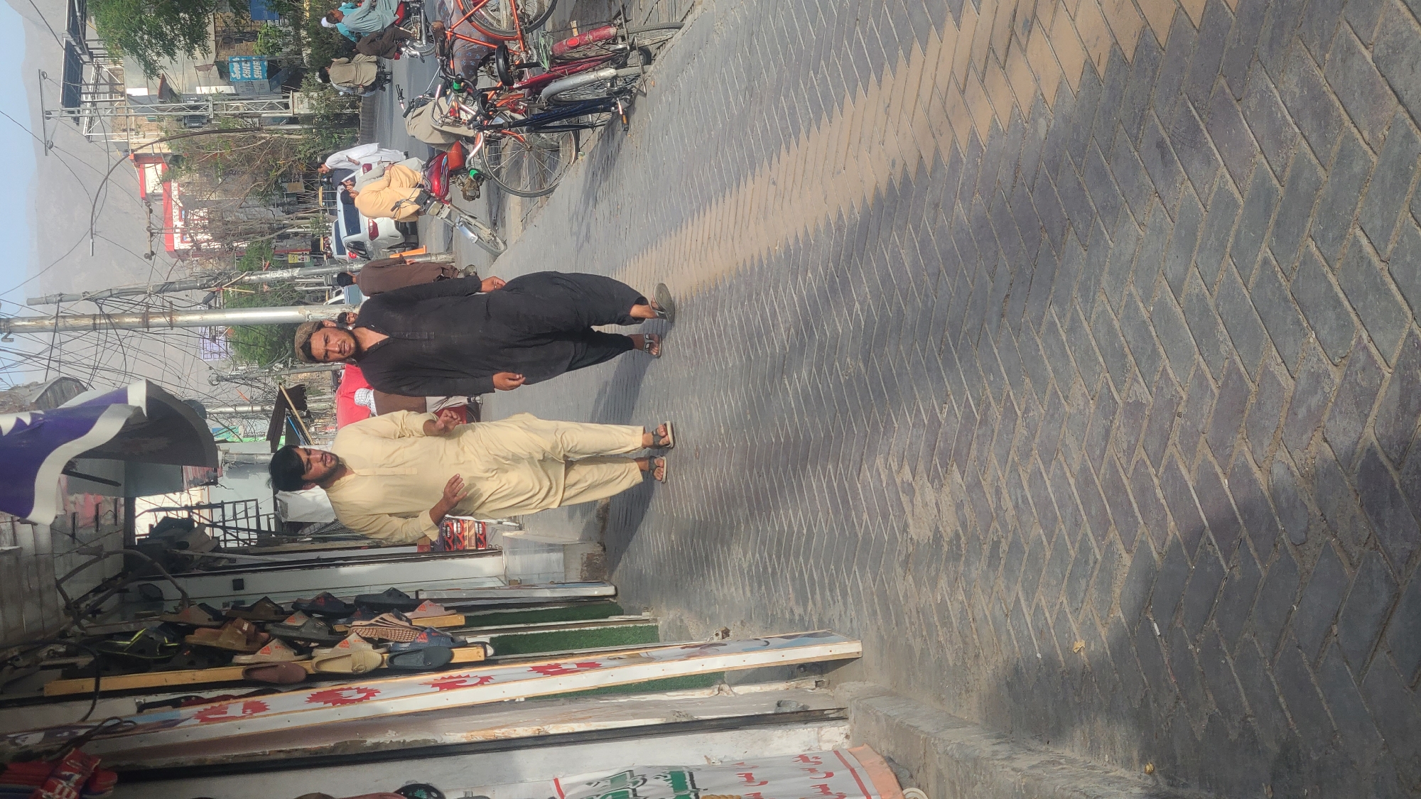 In Quetta City Tehsil, Pakistan, two men walk on a herringbone brick sidewalk next to a storefront. The man on the left wears a tan shalwar kameez, while the man on the right wears a black shalwar kameez and a cap. The storefront displays various footwear. Beyond the sidewalk, a street is visible with parked motorcycles and bicycles, as well as several cars and other pedestrians. Power lines crisscross above the street, and buildings and mountains can be seen in the background.