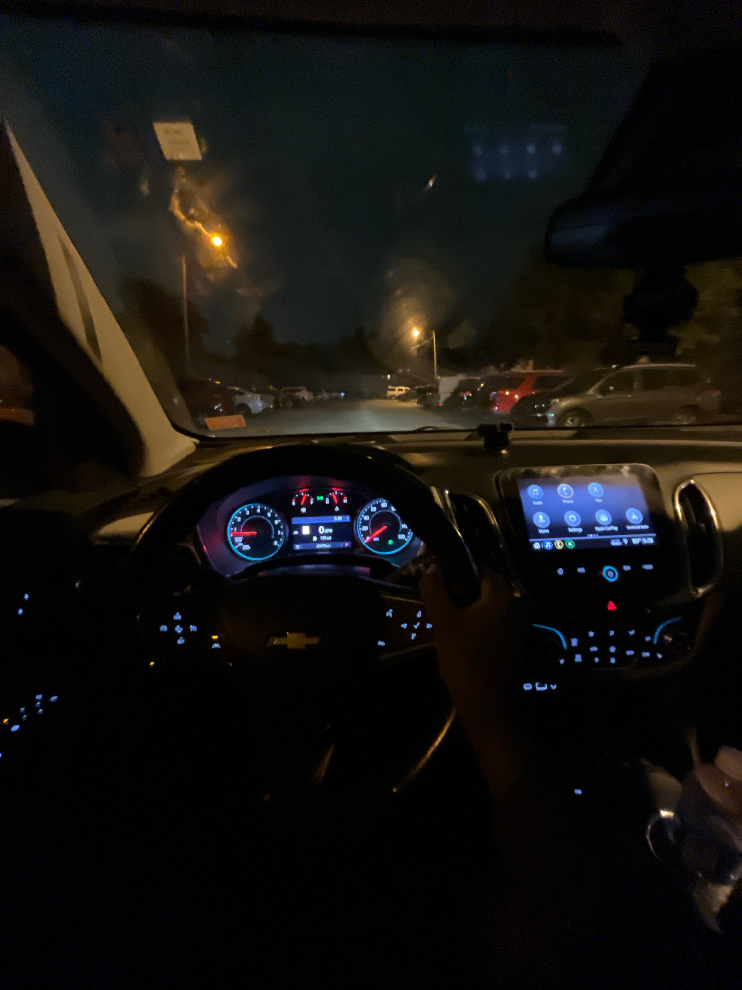 The image, taken at night, shows the interior of a car, likely in Louisville, United States of America. The driver's perspective is captured, with the steering wheel and dashboard prominently displayed. The dashboard features illuminated gauges, including a speedometer showing 0 MPH, and a central infotainment screen displaying various icons such as "Radio," "Phone," and "Apple CarPlay." Outside the car, streetlights and parked vehicles are visible, suggesting an urban or suburban setting. The scene is dark, with the primary illumination coming from the car's interior lights and the external streetlights.