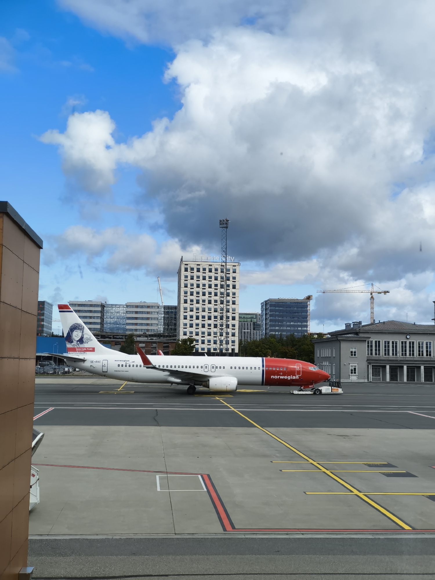 An exterior medium shot captures a Norwegian airline Boeing 737-800 aircraft, registered as SE-RXC, parked on an airport tarmac. The aircraft is predominantly white with a red nose and tail fin, featuring the "norwegian" logo and a portrait of a woman on its tail. A ground tug is attached to the front landing gear. The background includes several modern, glass-fronted buildings and a historical-looking grey building with columns, indicative of an airport terminal. Construction cranes are visible in the distance, suggesting ongoing development. The sky is partly cloudy with a mix of blue sky and large, grey clouds. The scene is likely at Tallinn Airport (TLL) in Tallinn, Estonia, given the architectural style and presence of Norwegian Air Shuttle.