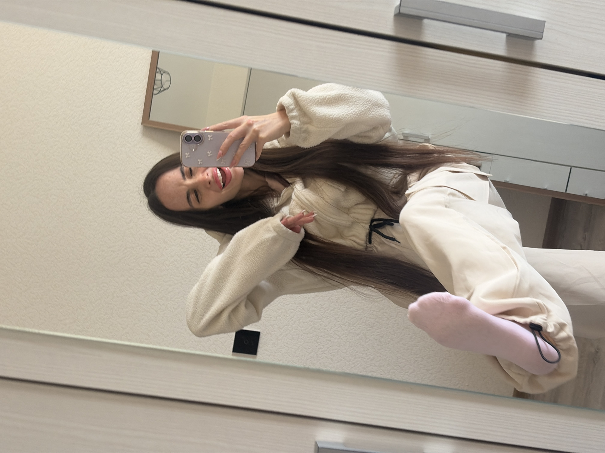 A young woman is depicted in a mirror selfie taken indoors. She holds a smartphone, encased in a light purple cover adorned with white embellishments, in her left hand, positioned to the right of her face. Her long, straight, dark brown hair flows over her shoulders. She is wearing a cream-colored, textured two-piece athletic or lounge set, featuring a hooded top with a quarter-zip and a cinched drawstring waist, paired with matching loose-fitting cargo pants that have elasticated cuffs and black drawstrings. Her right leg is lifted, revealing a light pink sock on her foot. Her facial expression shows an open mouth with her tongue slightly extended and partially closed eyes, indicative of a playful or cheerful demeanor.
The background, reflected in the mirror, consists of a textured, light beige wall. To the left of her reflected body, a small, square black object, possibly a light switch, is mounted on the wall. Above and to the right of her reflected head, a smaller, framed mirror with a wooden border is visible, reflecting a distinctive overhead light fixture with a basket-like or circular design. On the far right of the image, a segment of a light-colored wardrobe or cupboard with a prominent grey horizontal handle is visible, suggesting the mirror is integrated into or placed adjacent to this furniture. The floor is largely obscured, with only a sliver of light-colored flooring visible at the bottom left. There are no visual cues within the image that allow for the specific identification of a location within the city of Saint Petersburg, Russia.
