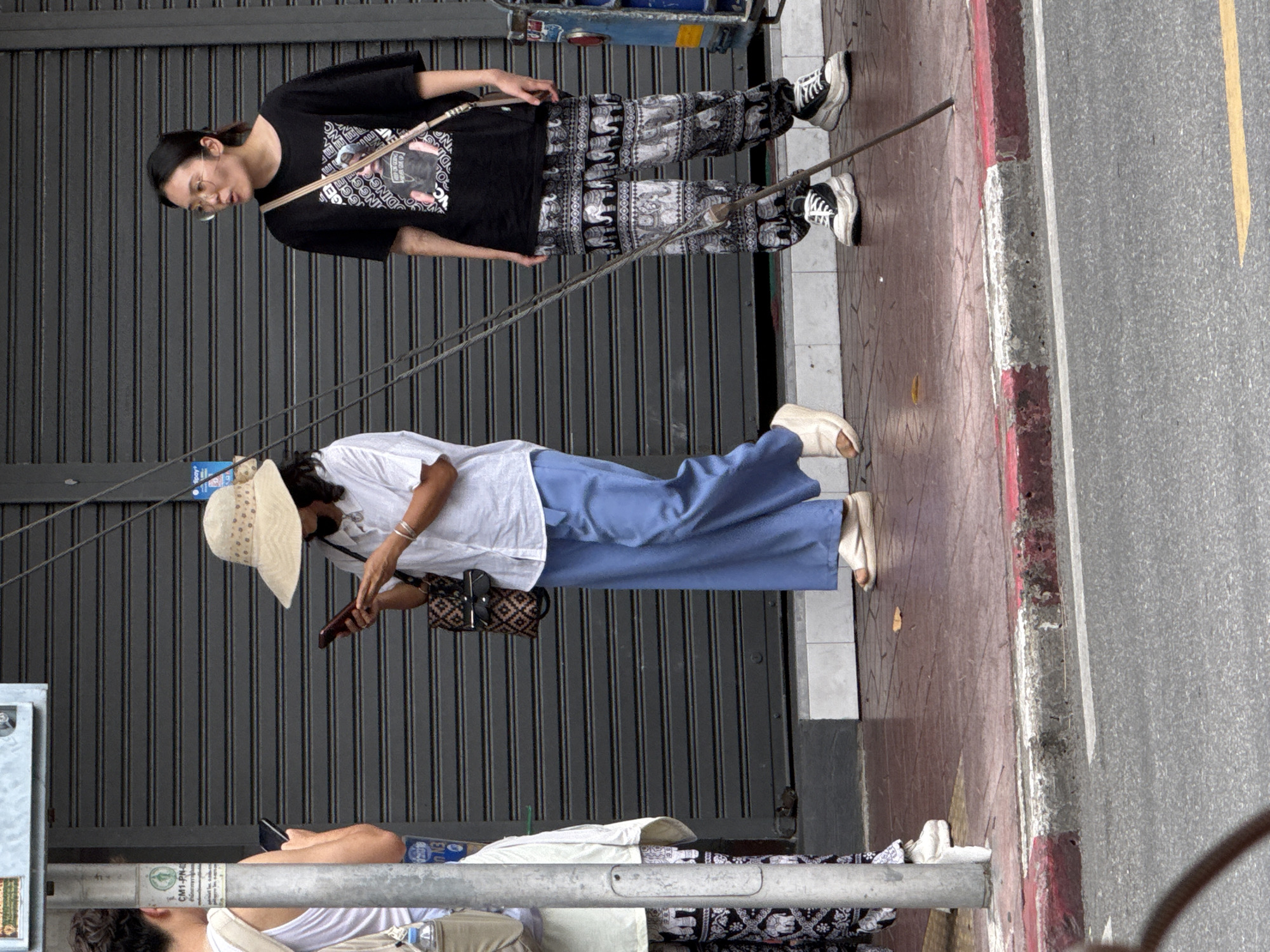 Three individuals are standing on a sidewalk in front of a grey metal shuttered storefront. The person on the left is partially visible from the back, wearing a white tank top and patterned pants. They are holding a dark-colored smartphone. In the center, a person wearing a wide-brimmed straw hat and a white shirt is looking at their smartphone. They are wearing blue wide-leg pants and light-colored sandals, and carrying a patterned tote bag. The person on the right is wearing glasses and a black t-shirt with a graphic print. They are wearing black and white patterned pants with an elephant motif and sneakers. This individual is looking to their left, with their mouth slightly open. A thin metal pole is stuck into the ground in front of them. The sidewalk is paved with reddish-brown tiles. The storefront behind them is closed. The image appears to have been taken in Bangkok, Thailand, given the common architectural style of the shutters and the general urban environment.