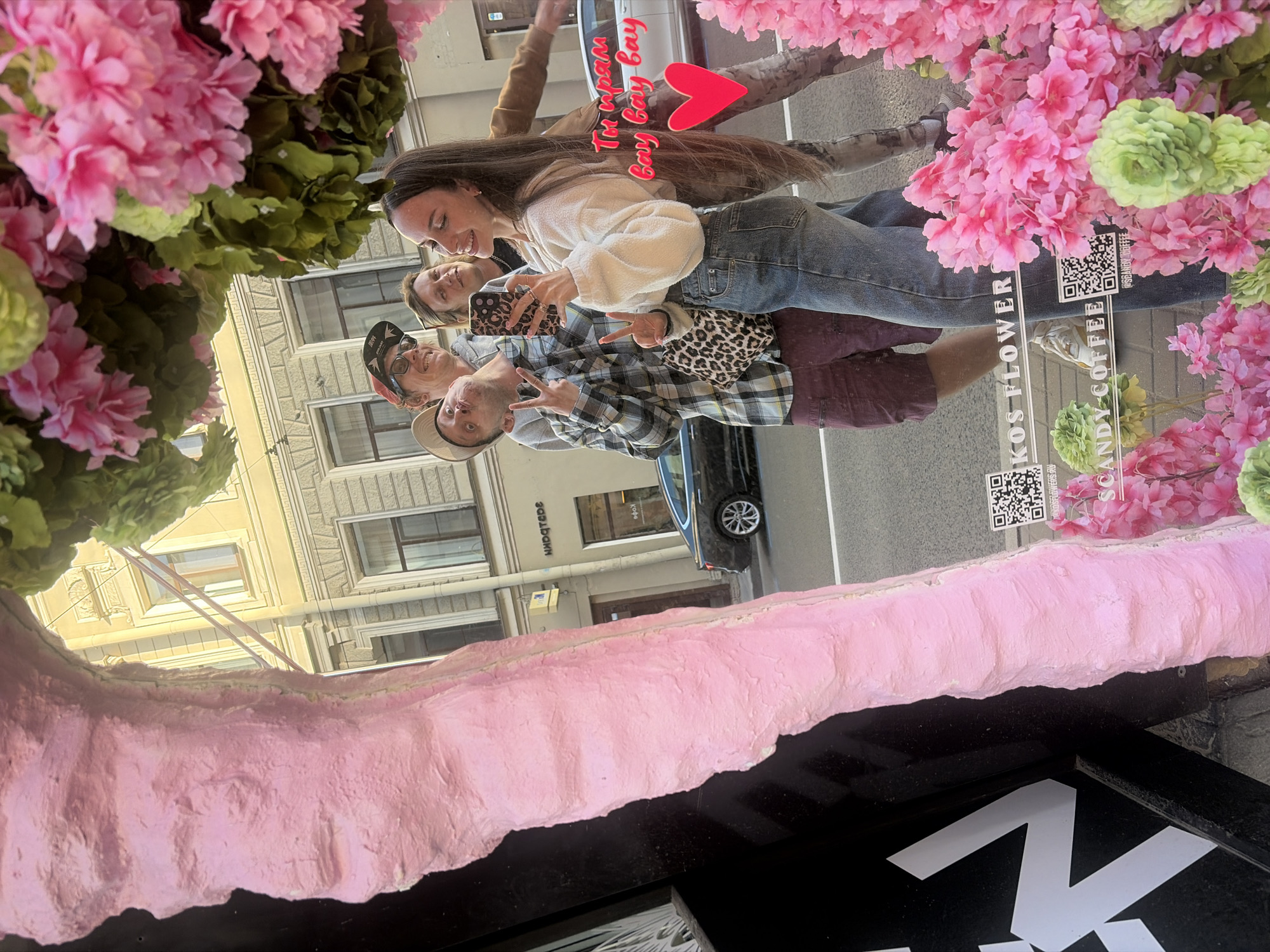 The image shows a group of four young adults reflected in a window display. From left to right, the first person is a male wearing a beige baseball cap and a plaid shirt, making a peace sign with his left hand and sticking out his tongue. Next to him is another male with blonde hair, wearing glasses and a grey hoodie, looking towards the right. A female with long dark hair is in the center, holding a smartphone with a leopard-print case and smiling. Behind her, another person with long brown hair is partially visible.
The background reflection shows a light-colored building with multiple windows and architectural details, typical of the historic center of Saint Petersburg. A black car is parked on the street in front of the building.
In the foreground, there's a large, textured pink object on the left, and a display of vibrant pink and green artificial flowers on the right. Text on the window display reads "KOS FLOWER" and "SCANDY COFFEE". A QR code is also visible on the window. There is handwritten text in pink and a red heart drawn on the reflection of the people, which appears to be added as a digital element. The overall scene suggests a casual outing and photo opportunity in a city environment.