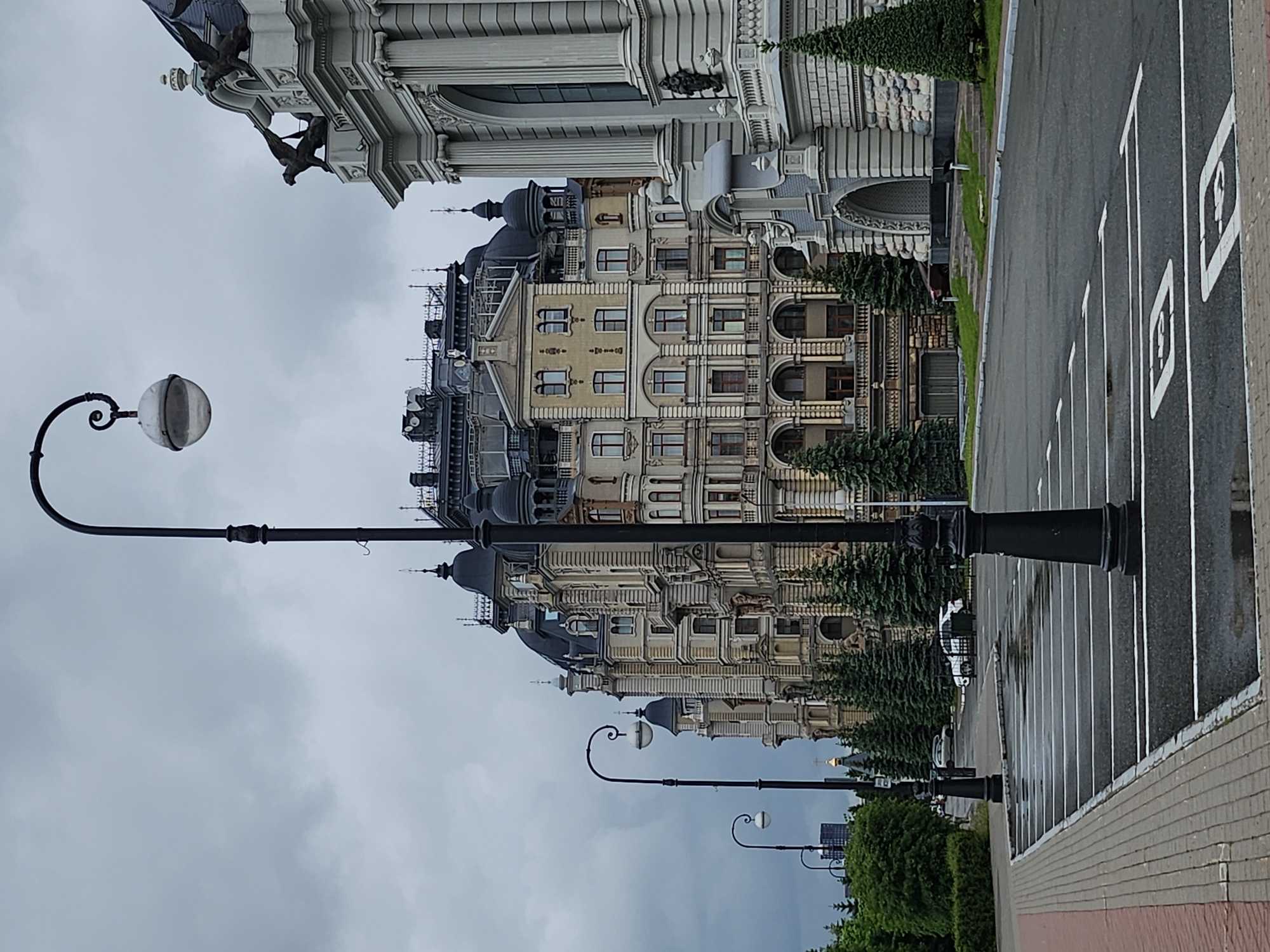 The image depicts an urban scene with a striking architectural presence. In the foreground are antique-style lampposts with globe-shaped lamps, positioned along a street with painted parking spaces. Beyond these, the focus is on a large, ornate building complex featuring a mix of architectural styles. The buildings have multiple stories with detailed facades, arched windows, and decorative elements like sculptures. Construction or maintenance work is visible on some parts of the structure. The sky is overcast, casting soft light on the scene. The location appears to be a city setting with well-maintained infrastructure and grand, historical architecture.