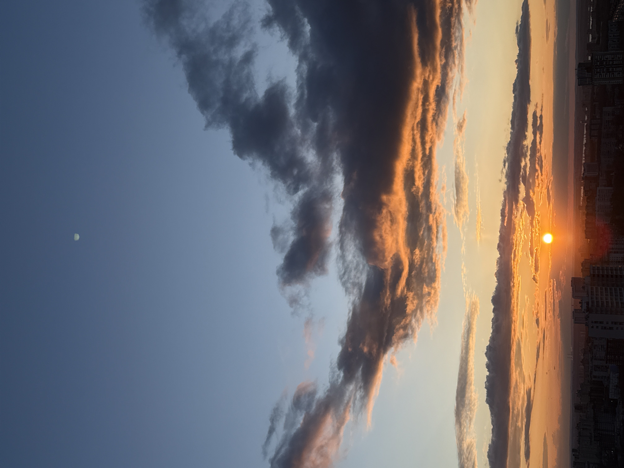 A breathtaking sunset paints the sky over a city, likely Perm, Russia, with vibrant hues of orange and gold. The low-lying sun, a brilliant golden sphere, is visible towards the bottom right of the frame, casting a warm, intense glow across the horizon and reflecting off the undersides of the clouds. Dramatic cloud formations stretch across the sky, with their dark, grey bodies contrasting sharply with their edges illuminated in fiery orange and deep gold by the setting sun. Above these illuminated clouds, the sky transitions to a serene blue, particularly in the upper left quadrant. A small, pale, circular object, likely the moon, is faintly visible in this blue expanse. Along the bottom edge of the image, the silhouetted skyline of a dense urban area, characterized by numerous buildings, stretches across the panorama. No people or visible text are present.