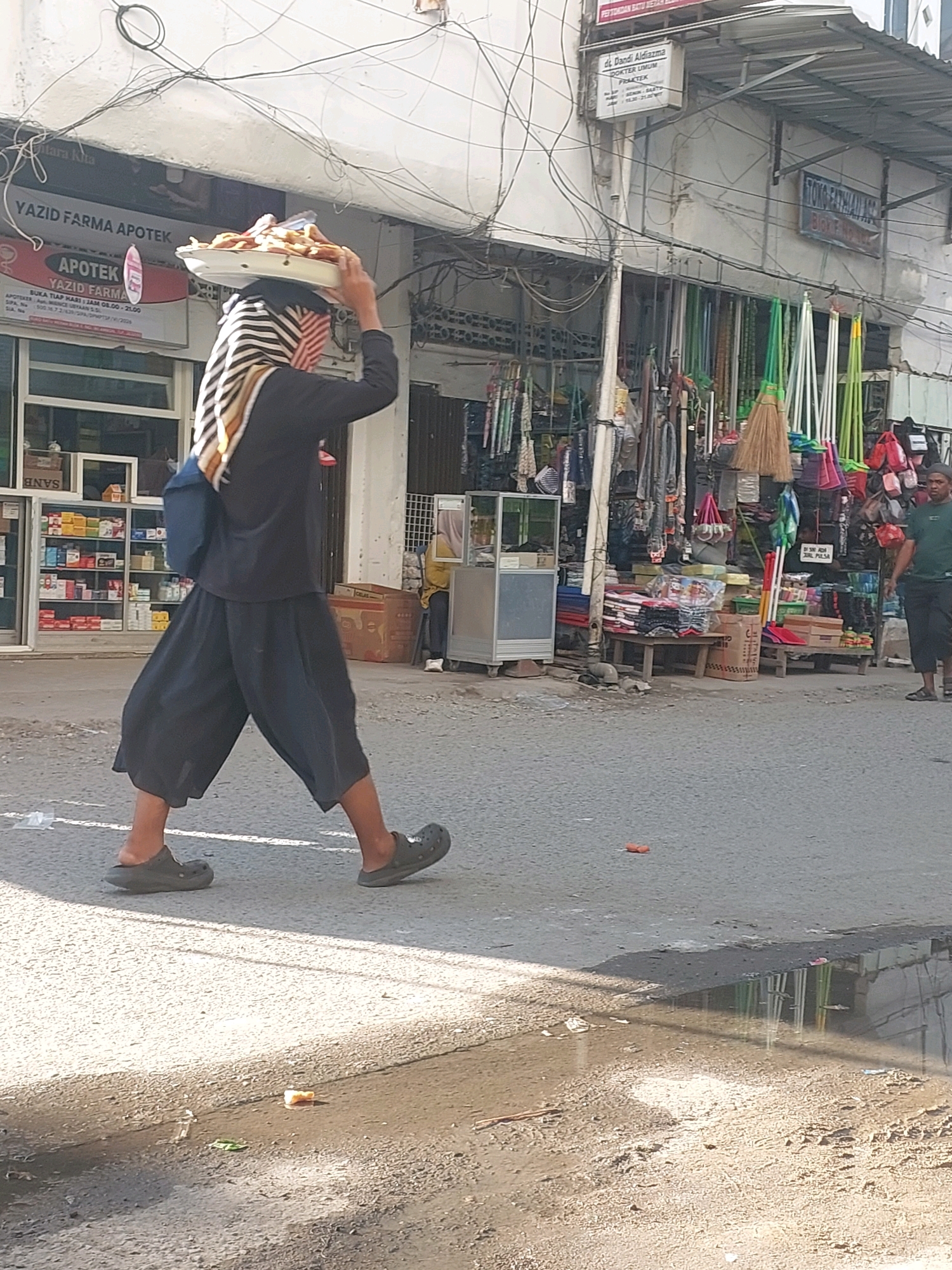 A person walks down a street in Ambon, Indonesia, carrying a tray of food on their head. The person is wearing black, loose-fitting pants and a black long-sleeved shirt. They are also wearing dark-colored clogs. The street is paved and appears to be wet in places, with puddles reflecting the surroundings. The person is walking past a row of shops that are selling various goods, including clothing, bags, and household items like brooms. The shops have awnings and signs, some of which are in Indonesian. The buildings are made of concrete and have exposed electrical wires running overhead. The scene appears to be a busy marketplace or shopping area.