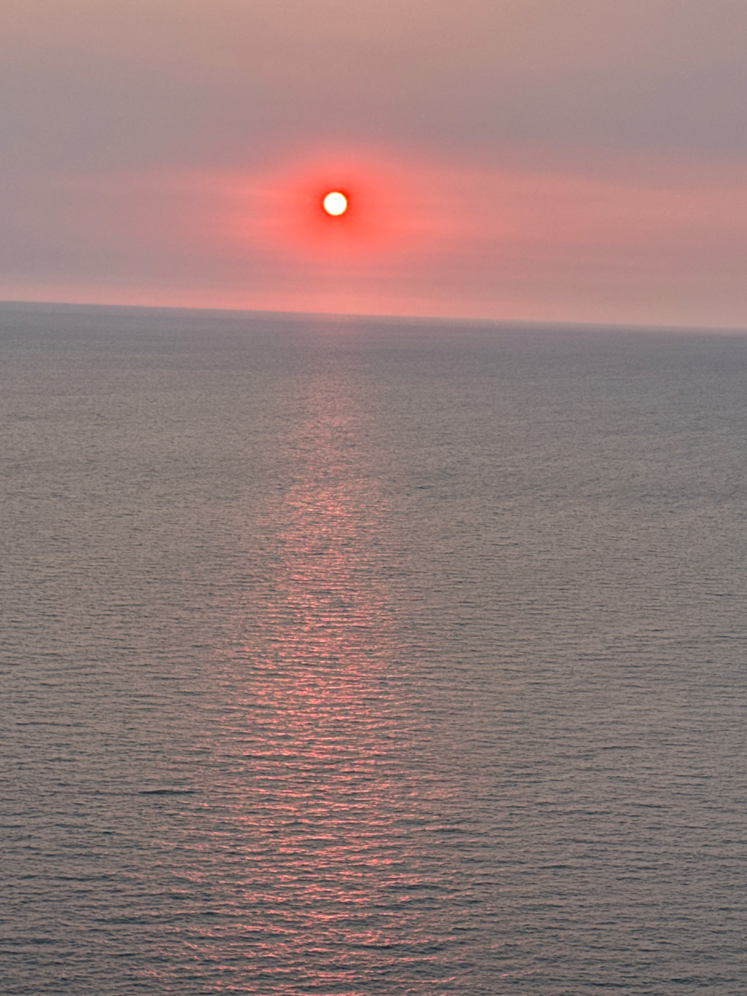 A serene sunset over the Adriatic Sea near Glavatičići, Montenegro, bathes the sky and water in a soft, rosy glow. The sun, a vibrant red orb with a bright white center, hovers just above the horizon, casting a shimmering path of light across the gently rippling water. The sea itself is a muted gray-blue, reflecting the diffused light of the sky. The horizon line is clear and distinct, separating the tranquil water from the hazy, pink-tinged sky. No people or structures are visible, creating a sense of vastness and solitude. The overall impression is one of peaceful natural beauty.