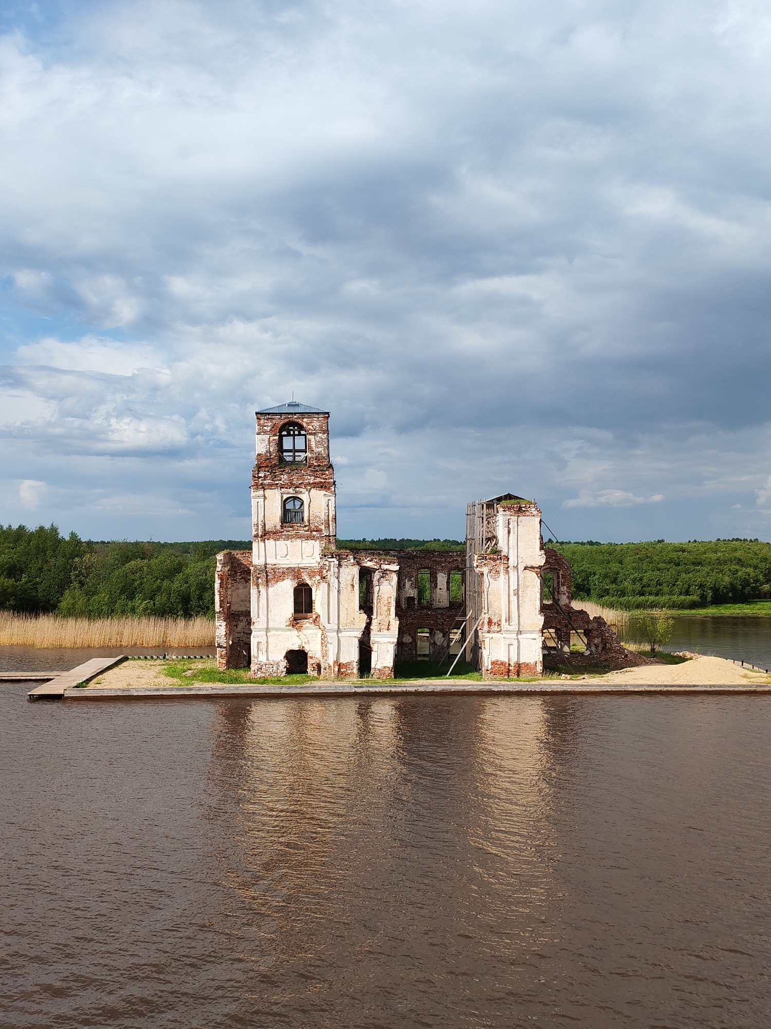 The image shows the ruins of the Church of the Nativity in Krokhino, Russia, standing on a small island in a body of water. The church is partially destroyed, with crumbling white and red brick walls, and a mostly intact bell tower. A wooden dock leads to the island. The water reflects the structure. In the background, there is a line of green trees along the distant shore. The sky is filled with grey and white clouds.