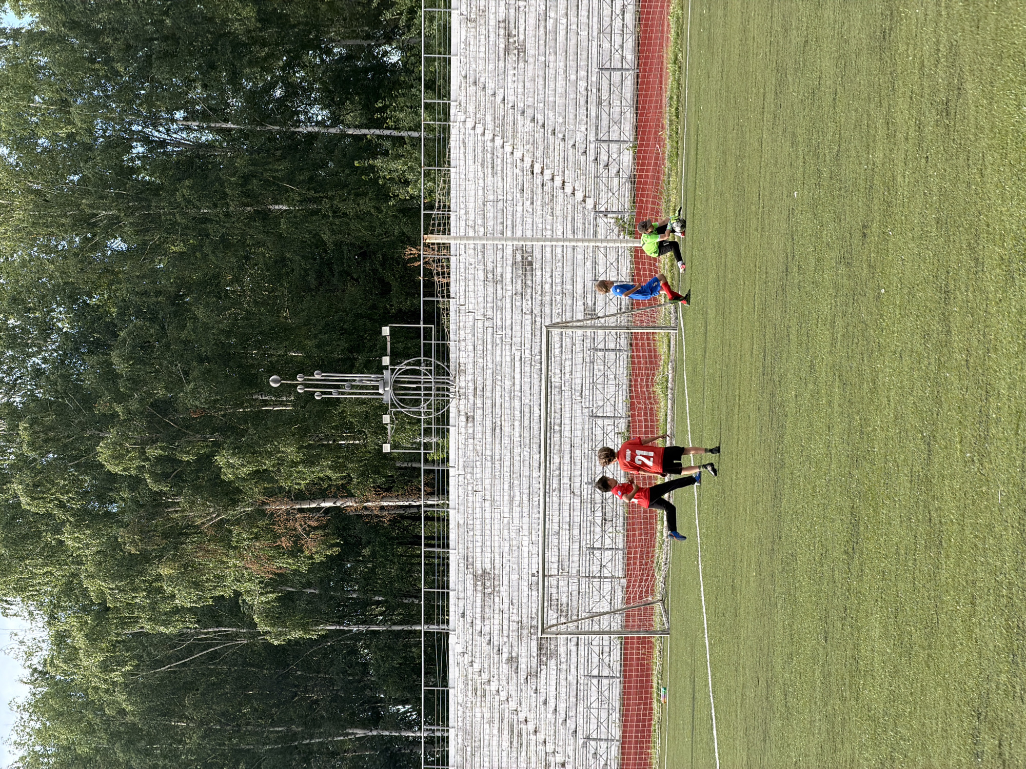 This media file depicts a sunny daytime scene on an outdoor artificial turf soccer field in Chaikovsky, Russia. The field is marked with white lines, and a soccer goal with a red net is visible. Beyond the field, a row of weathered, concrete stadium bleachers stands, with a dense line of green trees and birch trees behind them.
Several individuals are engaged in a soccer activity. Near the goal, a person in a bright green shirt and black shorts, likely a goalkeeper, is crouched and holding a soccer ball. To their left, a person in a blue shirt and red shorts is positioned as if ready to play. Further down the field, two individuals in red shirts and black shorts are interacting, one of whom is wearing the number "21" on their shirt. Their poses suggest they are actively participating in a game or practice.
The bright sunlight indicates it is likely mid-day. The lush green trees suggest a summer or late spring setting. The overall atmosphere appears to be one of energetic athletic activity in a somewhat rustic, natural environment.