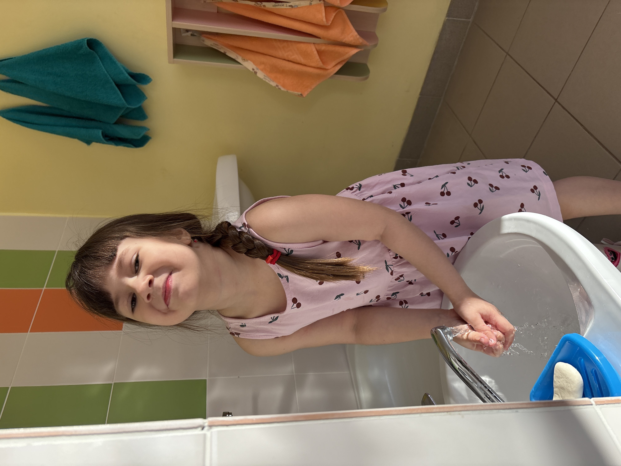 A young girl with brown hair and bangs, wearing a light pink dress with cherry patterns, is washing her hands in a white sink. Her hair is braided on one side and secured with a red tie. Water is running from the chrome faucet onto her hands. A blue soap dish with a white bar of soap is visible next to the sink. The bathroom has a yellow wall and tiled walls with horizontal stripes of white, orange, and green. Turquoise towels hang on the wall to the left, while folded orange towels are on a shelf above. The location is in the city of Chaikovsky, Russia.