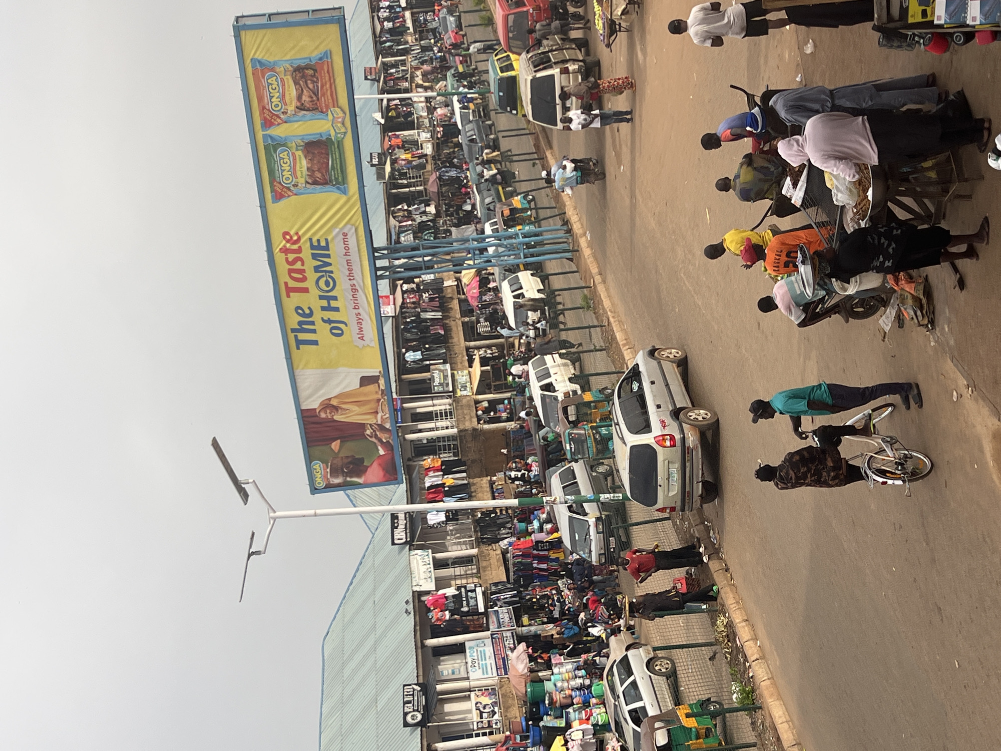 The media file captures a bustling street scene in Bukuru, Nigeria. A prominent billboard on the left advertises "The Taste of HOME," featuring images of Onga products, specifically beef stew and seasoning. The billboard's text also states, "Always brings them home."
The scene is characterized by a variety of people engaged in daily activities, likely commerce, given the presence of market stalls and various vehicles. Numerous individuals are seen walking, standing, or interacting. Objects include several vehicles, such as cars, a tuk-tuk, and a motorcycle, as well as market goods displayed for sale, such as clothing and other items in plastic bags. The setting appears to be an outdoor market or a busy street with shops lining one side.
The time of day is likely daytime, with ample natural light illuminating the scene. The weather appears to be clear. The overall atmosphere is one of lively activity and commerce, with a sense of community engagement. The people are dressed in casual attire, suggesting a typical day in a bustling urban or semi-urban environment.
Visible text includes brand names like "ONGA" and "PayPOS," as well as what appears to be store signage. The overall impression is of a vibrant and active marketplace in Nigeria.
