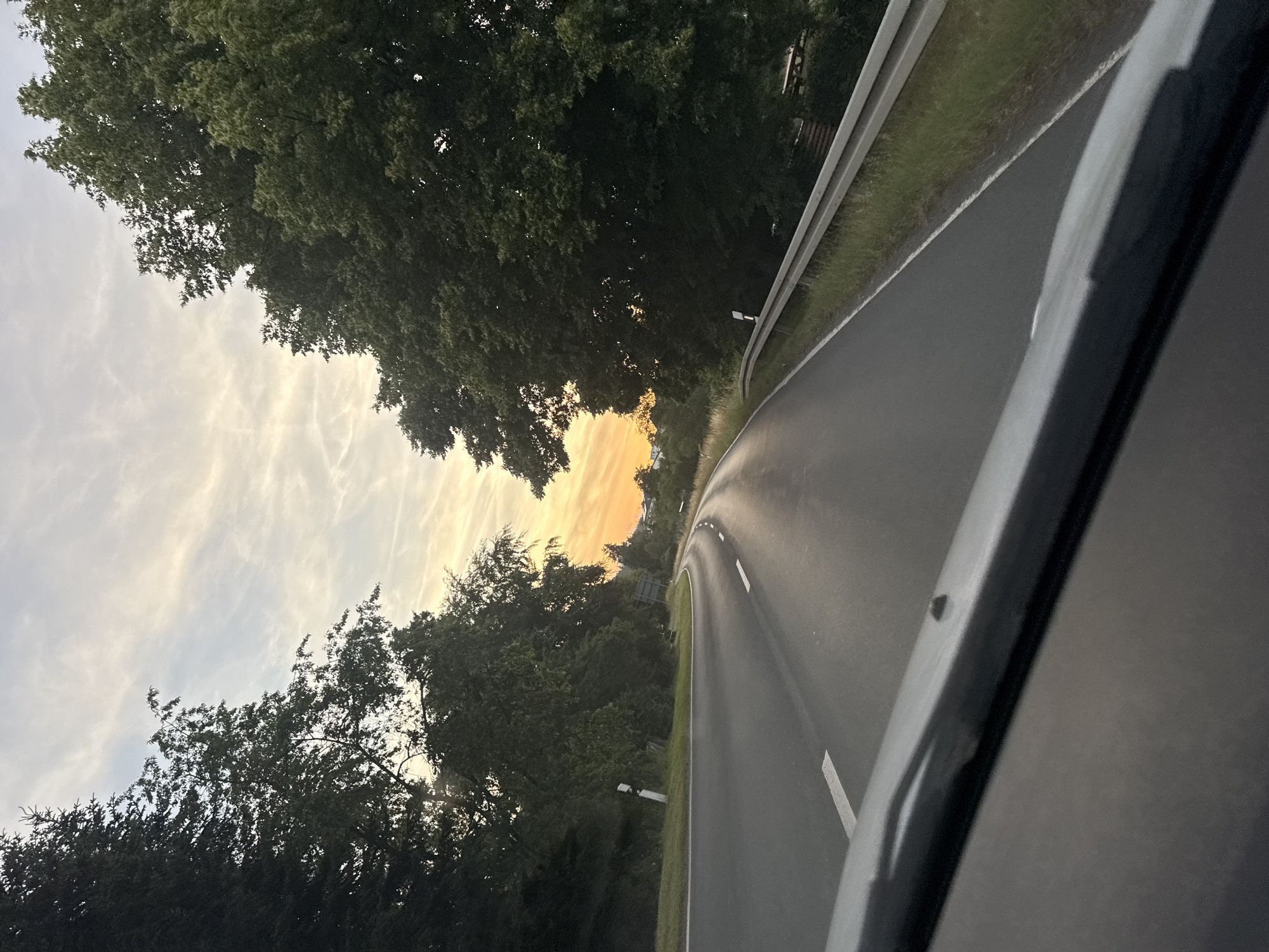 The image captures a scenic view of a road curving through a forested area during sunset. The perspective is from inside a vehicle, looking out. The road, paved and with white lane markings, dominates the right side of the frame and recedes into the distance. The left side of the image is filled with lush green trees, their dense foliage silhouetted against a dramatic sky. The sky itself is a spectacle of color, transitioning from a pale blue at the top to vibrant oranges and yellows near the horizon, illuminated by the setting sun. Wispy clouds streak across the sky, catching the warm light and adding texture.
There are no people visible in the image. The prominent objects include the road, the trees, and a metal guardrail running alongside the road. A few white road markers are visible along the roadside. The overall mood is tranquil and serene, enhanced by the natural beauty of the landscape and the warm glow of the sunset. The presence of the road and guardrail suggests a public road, possibly a rural highway. The time of day is clearly sunset, indicated by the low angle of the sun and the rich colors in the sky. The weather appears clear, with the clouds contributing to the picturesque sky rather than obscuring the sun. There is no visible text in the image.