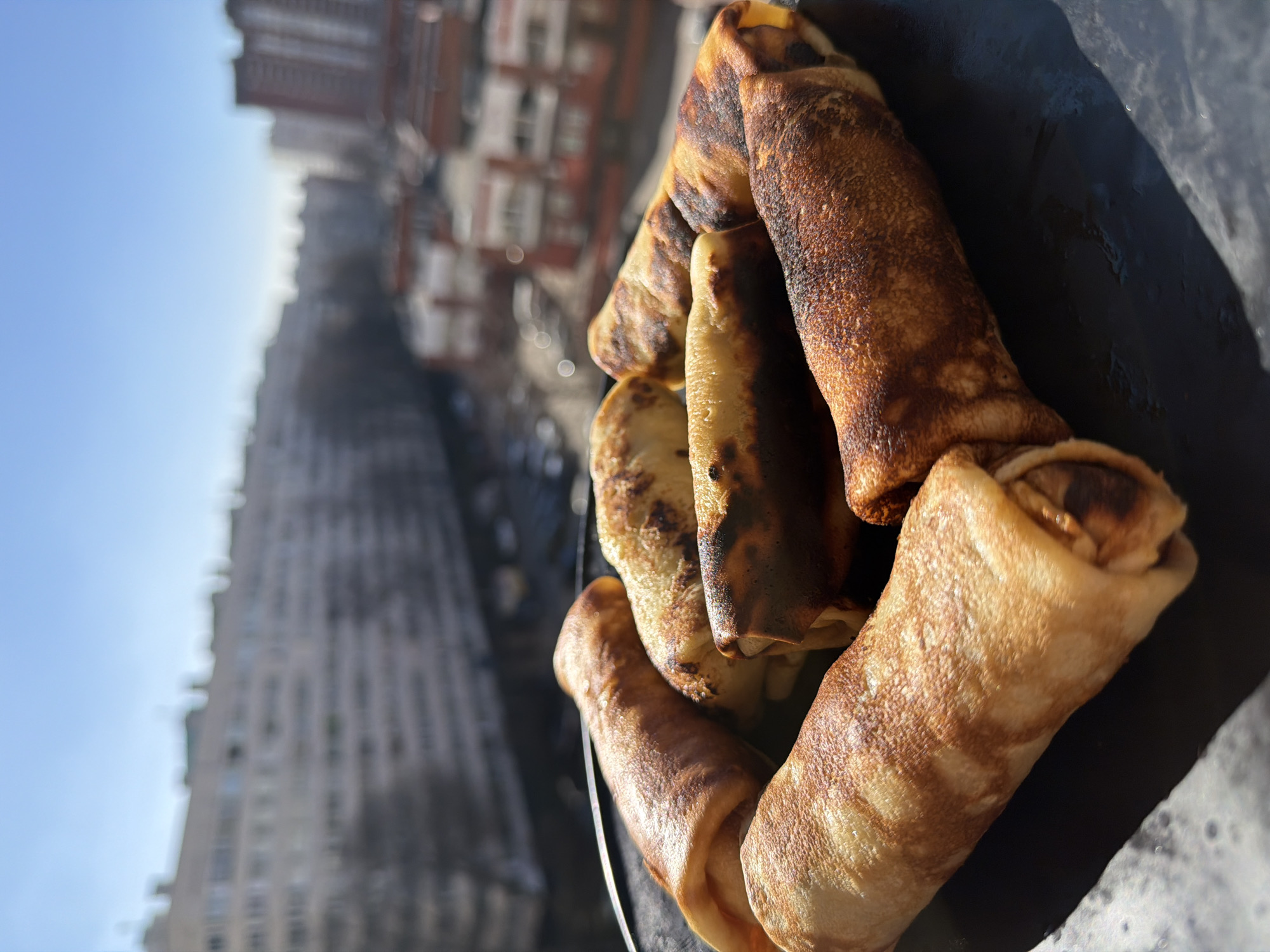 The image shows a plate of rolled pancakes, possibly blini, with an urban backdrop. The pancakes are arranged on a dark-colored plate, exhibiting varying degrees of browning, suggesting different cooking times or heat exposure. In the background, a cityscape is visible, dominated by tall buildings. The architectural style and urban layout hint towards the location being Saint Petersburg, Russia. The blurred background indicates a focus on the pancakes, emphasizing them as the primary subject. The image composition suggests a lifestyle or food photography context.