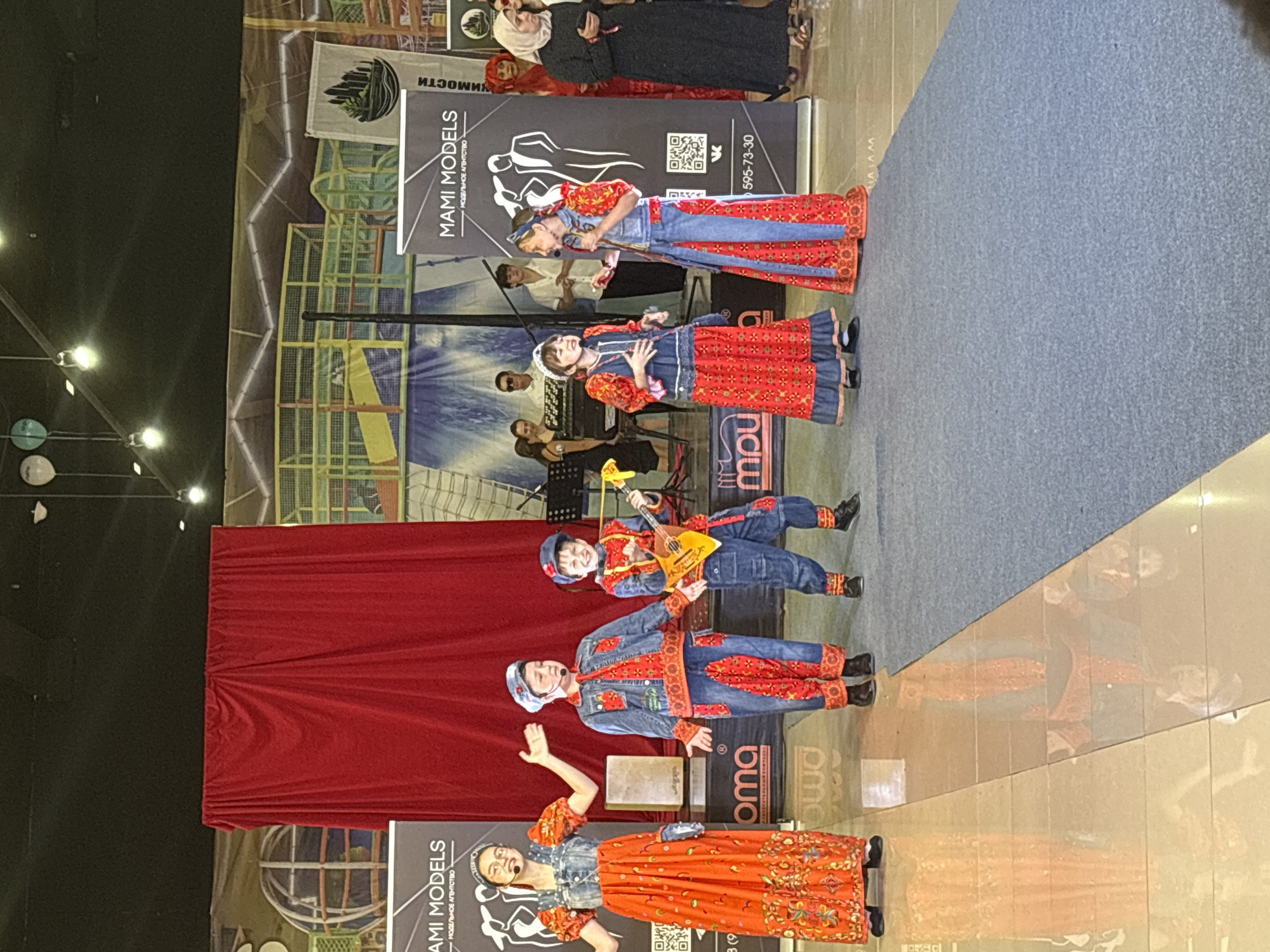 The image captures a stage performance featuring five children dressed in traditional-inspired costumes. The children, four boys and one girl, are arranged in a line, with the girl on the far left and two boys on the far right. The child in the center right is playing a balalaika. The stage has a red curtain in the background, with banners and a backdrop depicting a landscape. The floor is a reflective surface that mirrors the subjects. The scene appears to be indoors, possibly at an event or competition, given the presence of banners and audience members in the background. The overall atmosphere suggests a lively cultural performance.