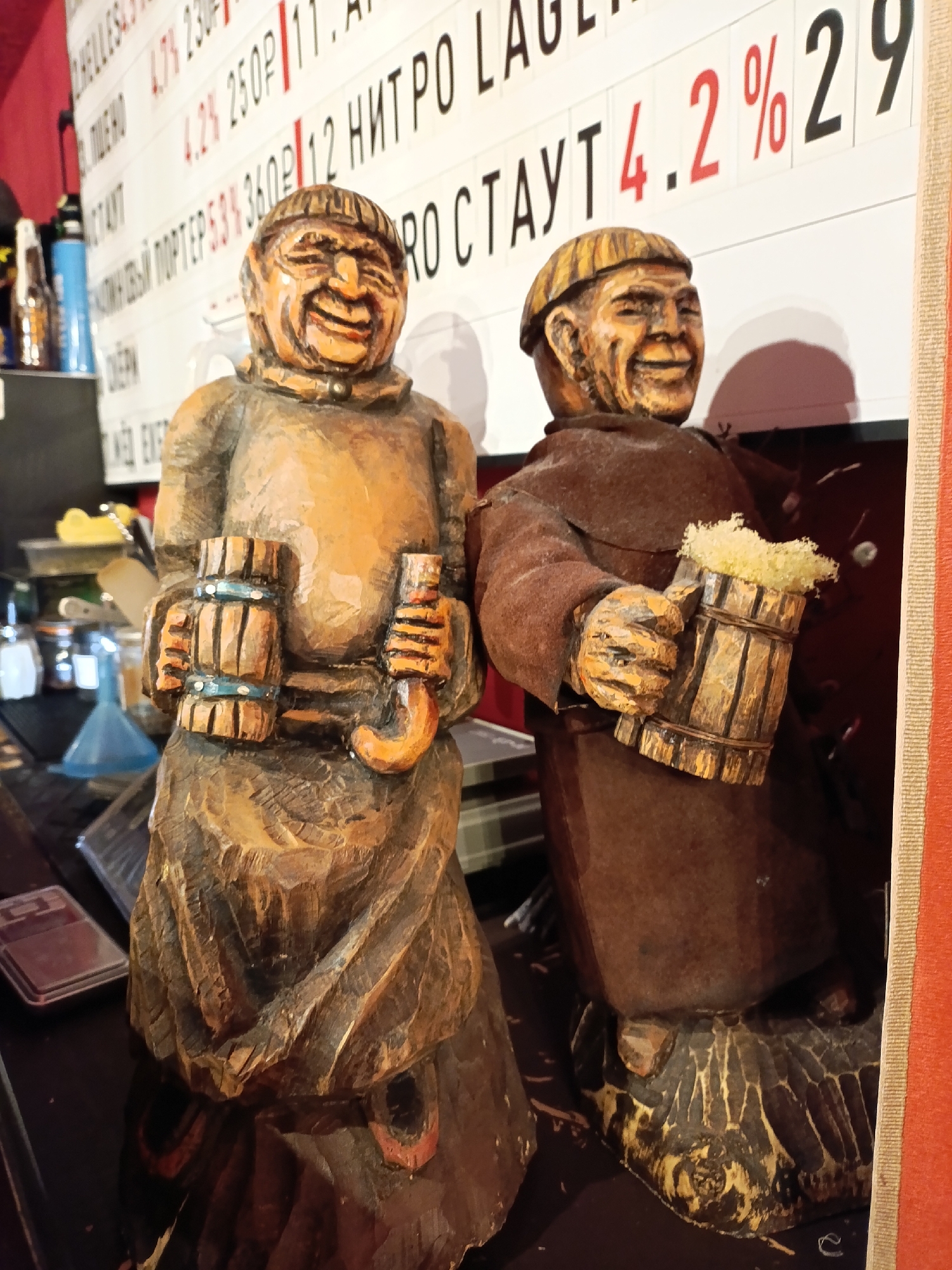 This media file shows two wooden figurines of men dressed as monks, holding mugs of beer. They are positioned on a dark surface, likely a bar top or shelf, in front of a white board with black and red text. The figurines are carved with detailed expressions and clothing, and the mugs appear to be filled with foamy liquid.
The text on the board suggests a bar or pub setting, possibly a menu, with prices and beer descriptions in Russian. Some visible text includes "4.2%", "250g", "12", "NITRO", and "PORTER". The overall mood of the figurines is jovial, with wide smiles and a celebratory feel, enhanced by the beer mugs.
The background shows a blurry assortment of objects, including bottles and jars, further indicating a commercial establishment. The lighting is somewhat dim, with a warm tone, suggesting an indoor environment. The context provided indicates the location is Chaikovsky, Russia. The scene captures a moment of apparent festivity or advertisement for beer.