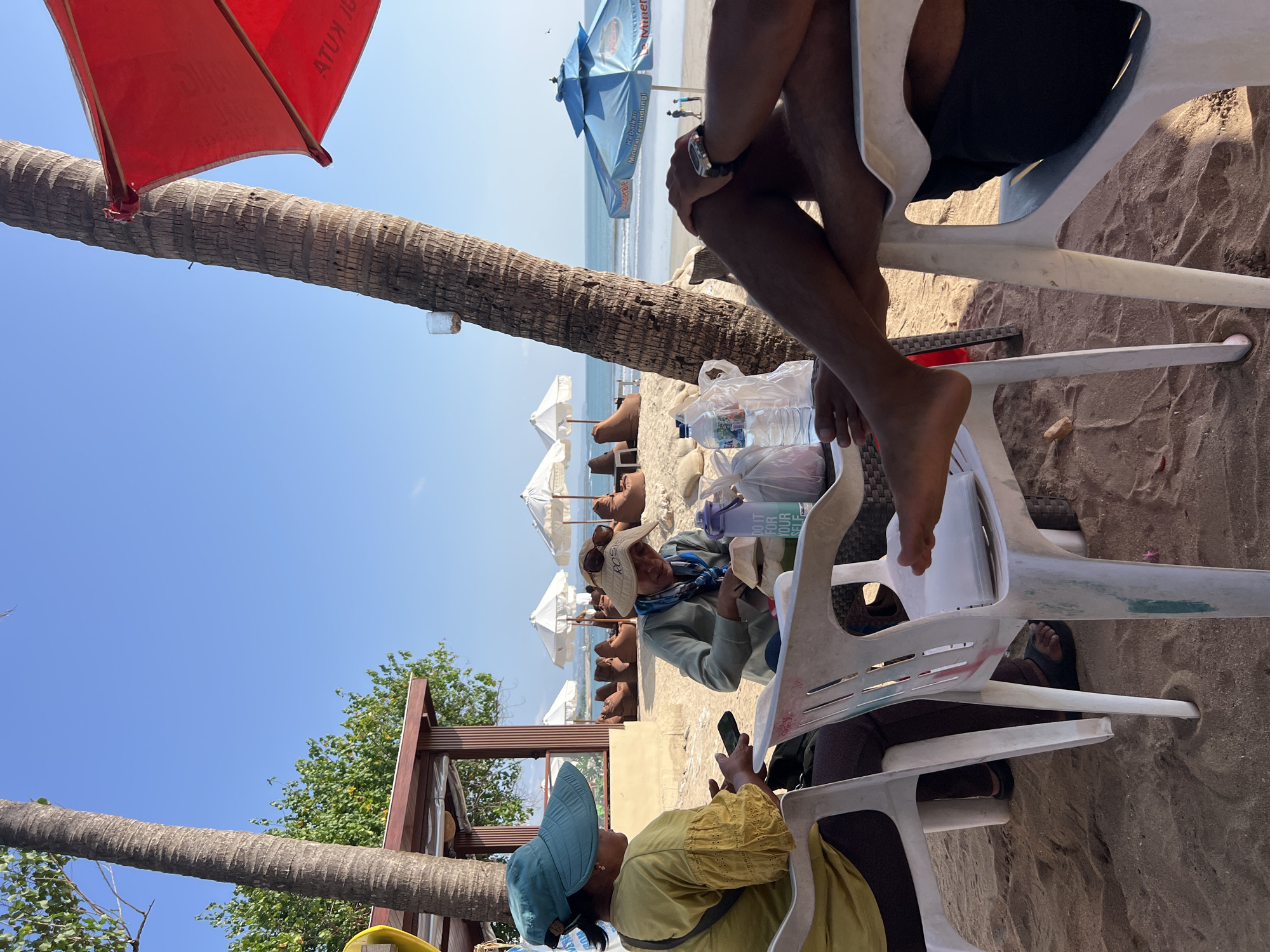 The image depicts a beach scene in Kuta, Indonesia, under a clear blue sky. Two individuals are seated at a white plastic table on the sand, likely enjoying a meal or drinks. One person, wearing a teal sun hat and a yellow top, is holding a mobile phone. The other individual, wearing a straw hat and a light blue shirt, is attending to a halved coconut. Several water bottles and plastic bags are also visible on the table. In the background, beach umbrellas, lounge chairs, and palm trees are scattered across the sandy shore. The ocean is visible beyond the beach, with gentle waves. A red umbrella is partially visible in the upper right corner.