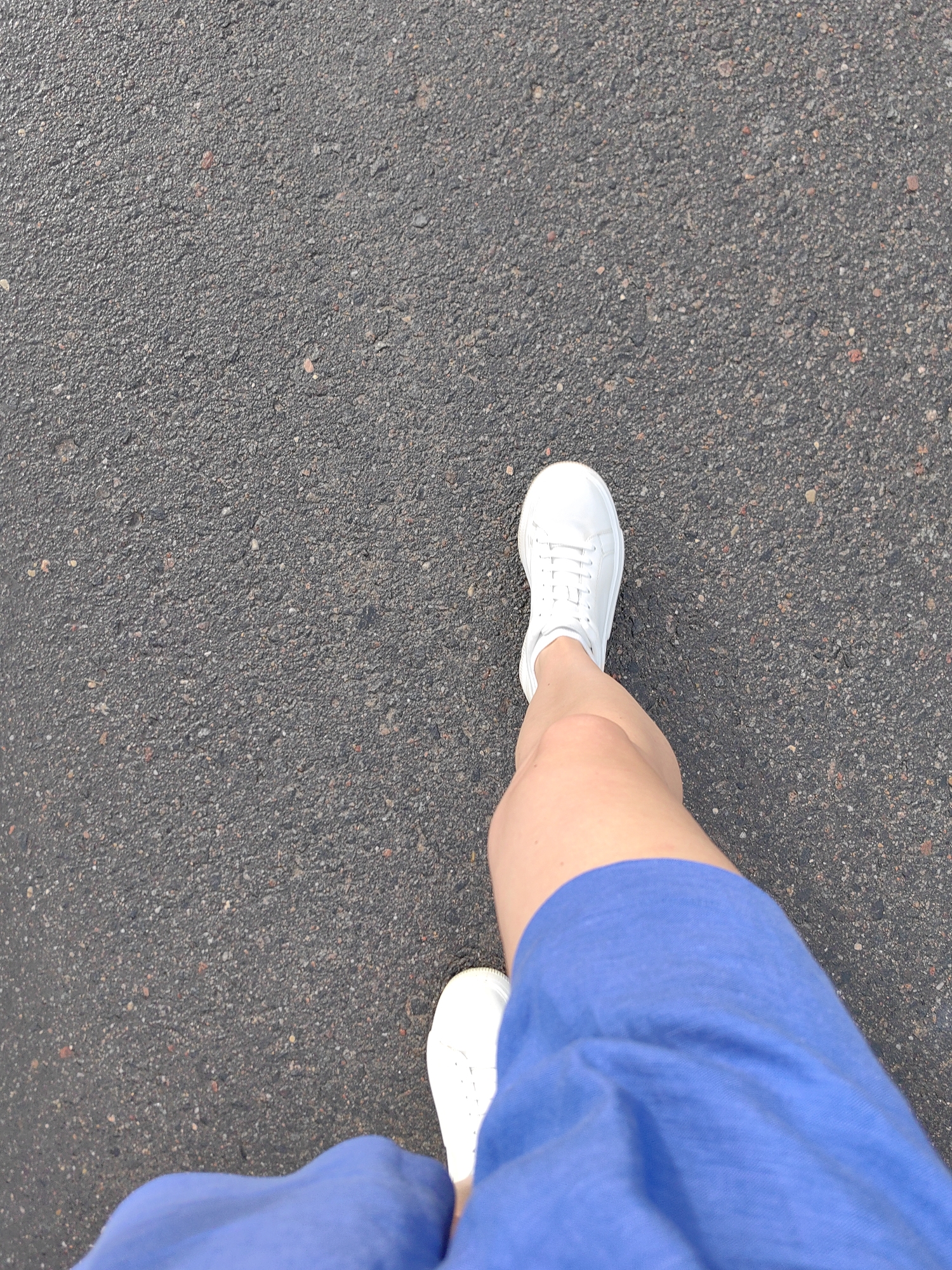 A first-person perspective shot captures a person walking on a paved surface. The person is wearing white sneakers and a blue skirt or shorts. The paving appears to be asphalt, with small stones and a dark gray color. The lighting suggests it is daytime. The scene is set in the city of Fanipal, Belarus.