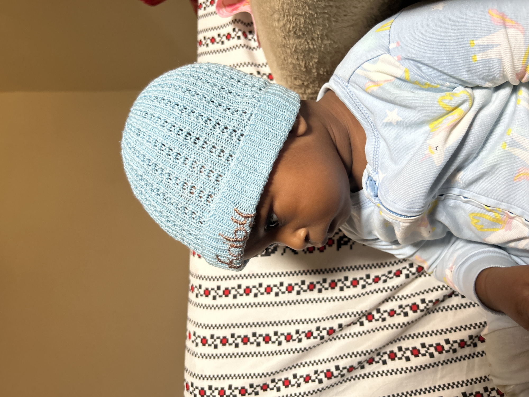 A young child is lying down, wearing a light blue knitted hat and light blue pajamas with unicorn and star patterns. The child's head is resting on a striped blanket with a pattern of red squares and black zigzags. The scene appears to be indoors, possibly a bedroom, given the soft lighting and bedding. The child is looking downwards and appears calm or sleepy. The image is taken from a slightly elevated angle, looking down at the child. The backdrop is a plain, light brown wall. No text is visible in the image. The location context is Jos, Nigeria.