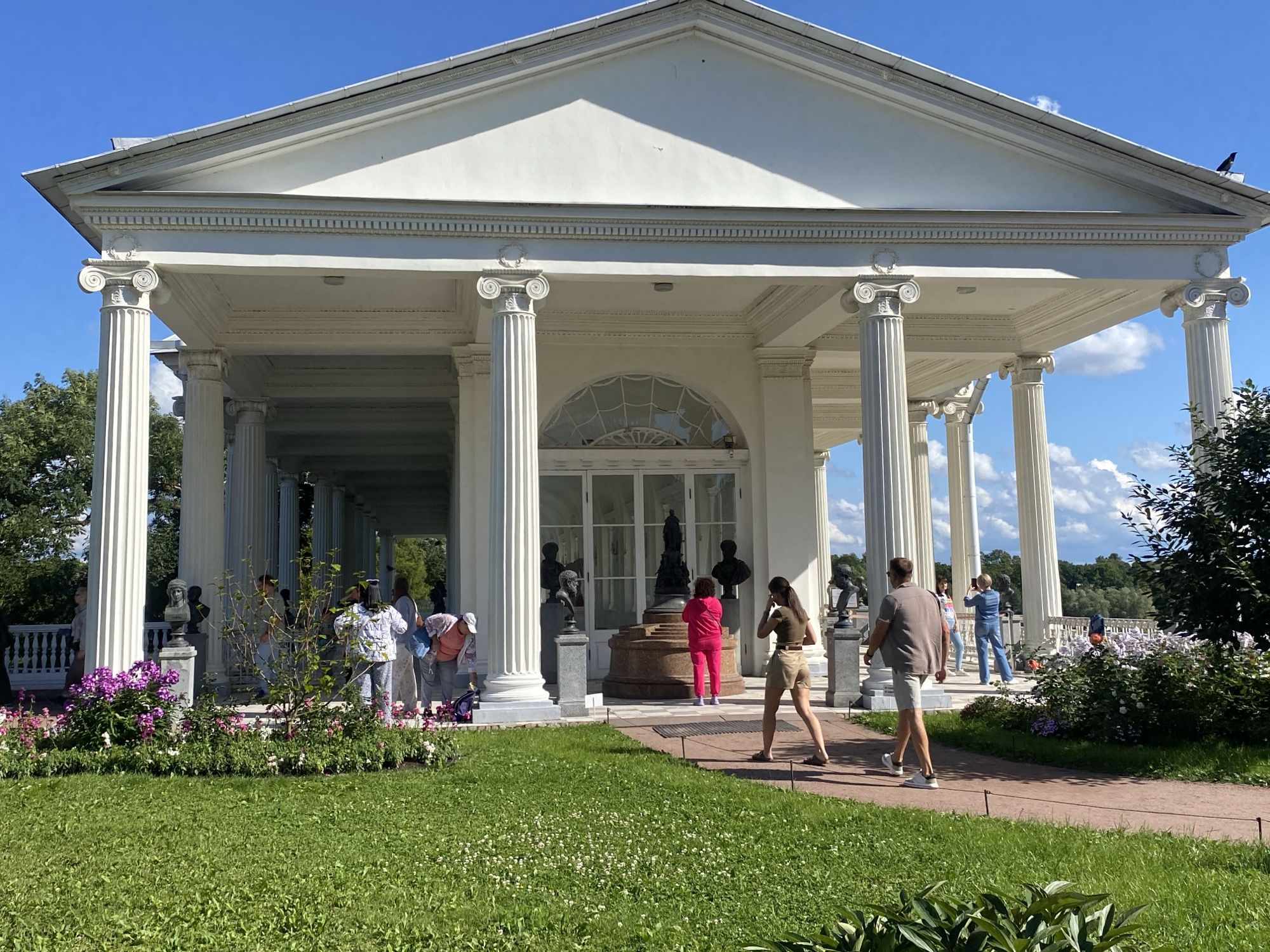 The image depicts a neoclassical pavilion with a portico supported by Corinthian columns. The pavilion is situated in a park setting with lush green grass, flowerbeds, and trees under a clear blue sky with scattered clouds. Several individuals are present, some walking along a path, while others are near the pavilion, observing statues and the architecture. One person is taking a photo of the pavilion. The pavilion appears to be part of the Catherine Palace complex in Pushkin, Russia, as evidenced by its architectural style and the parkland visible. The scene conveys a sense of leisure and historical appreciation in an outdoor environment.